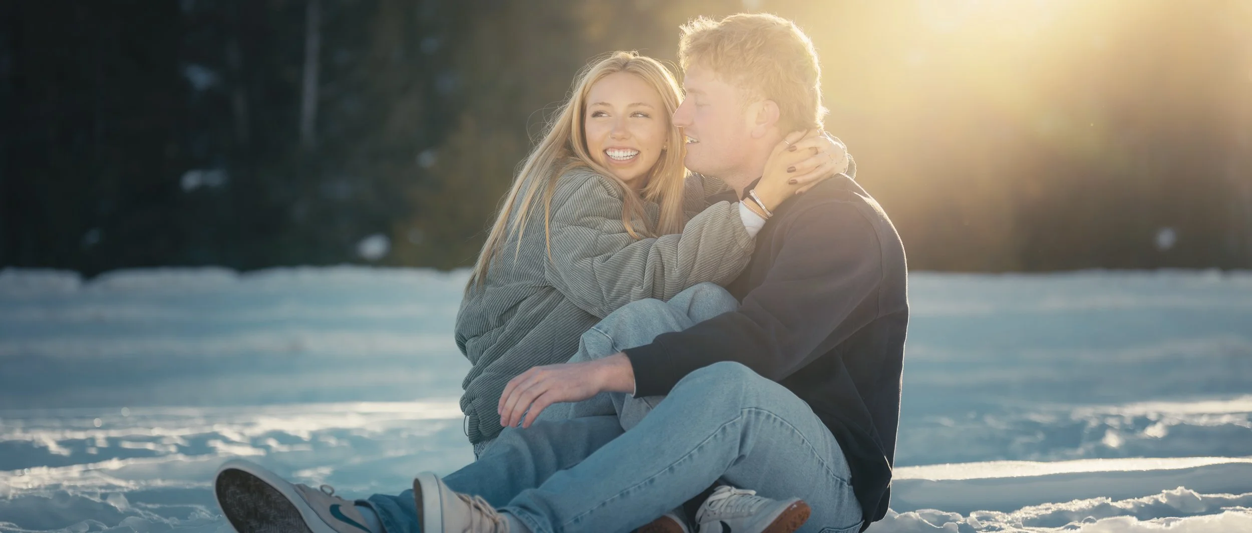 A young couple sitting in the snow during sunset, smiling and embracing each other.
