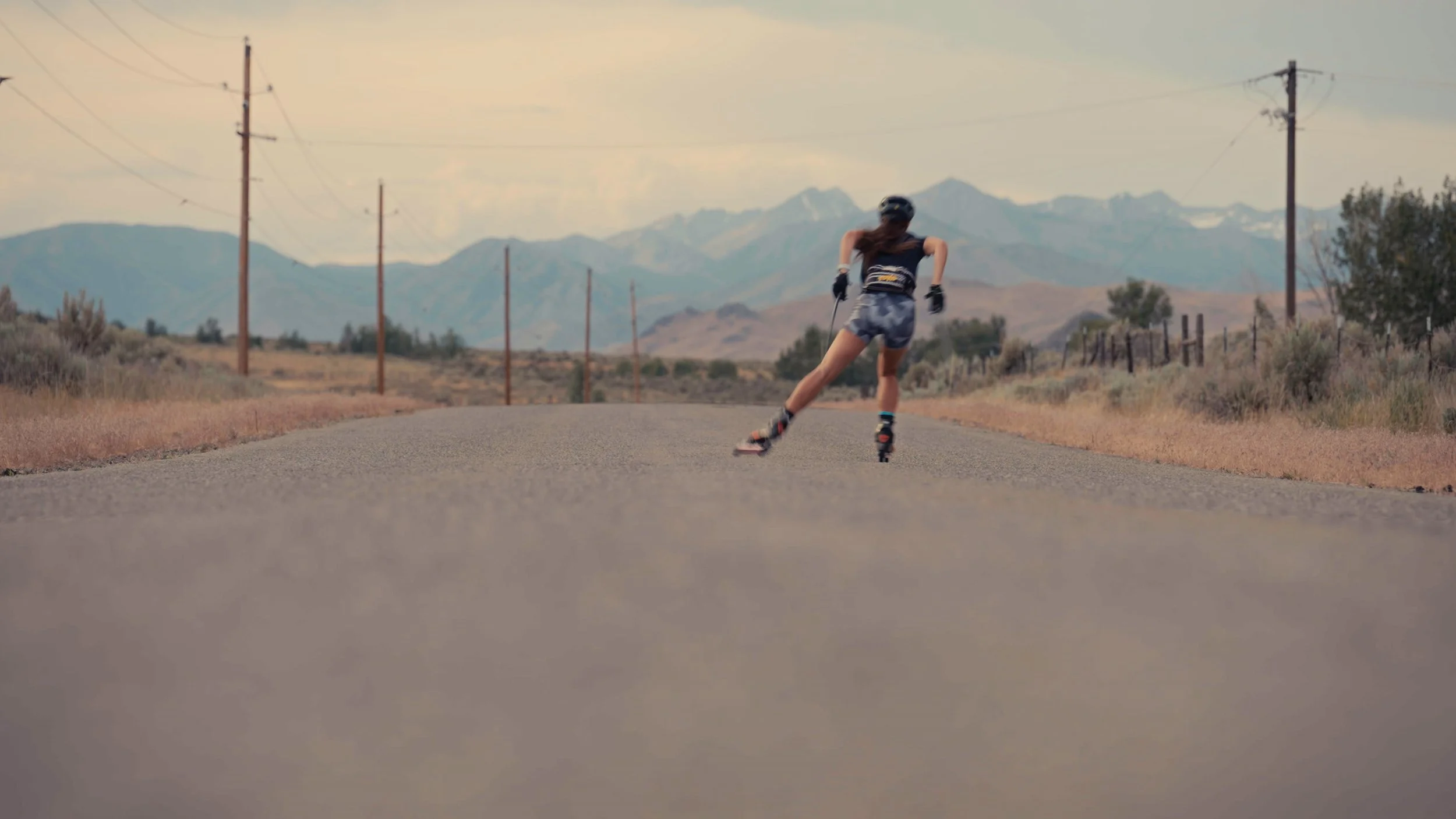 A person roller skiing on a paved road in a rural area with mountains in the background, wearing a helmet, sunglasses, and athletic clothing.