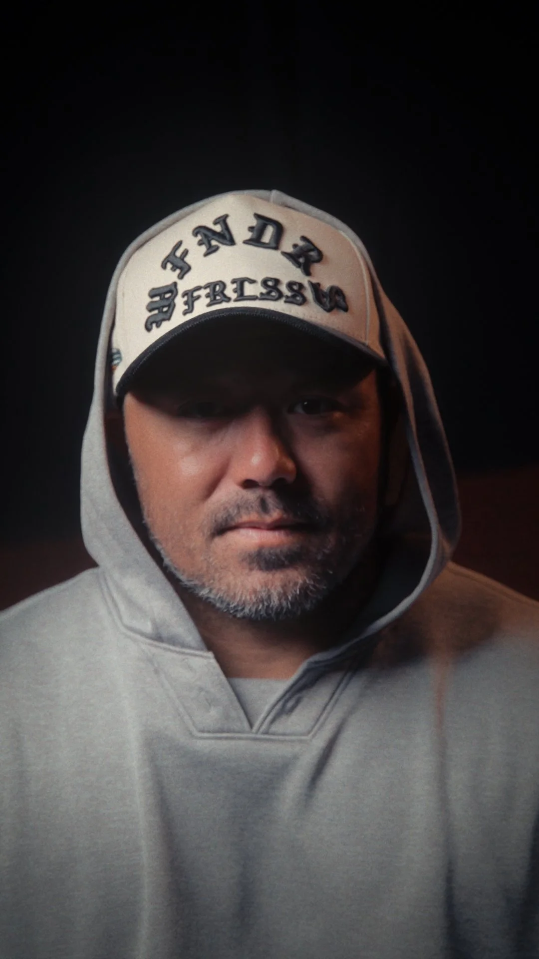 A close-up of a man wearing a gray hoodie and a baseball cap with the words 'FINDERS' on it. The background is dark.