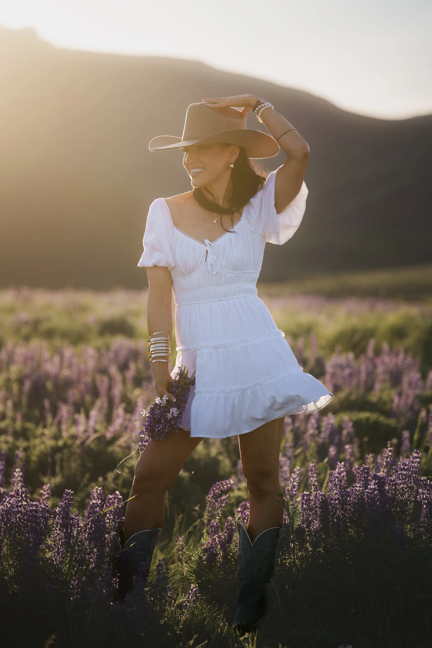 A woman in a white dress and cowboy boots standing in a field of purple flowers, wearing a large cowboy hat and smiling at sunset.