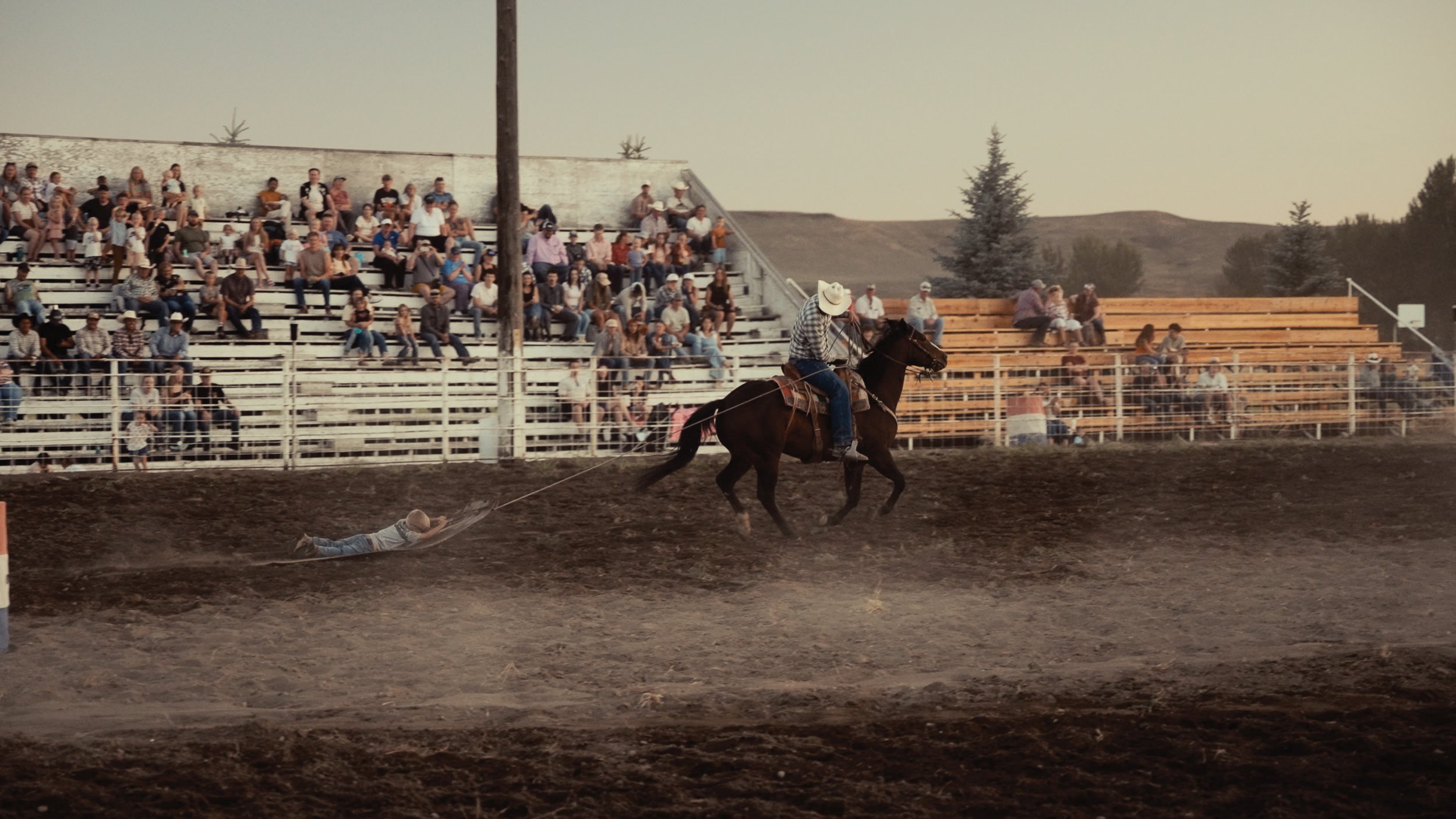A rodeo scene with a cowboy on a horse pulling a person on a rope behind them. Spectators are watching from bleachers in the background.