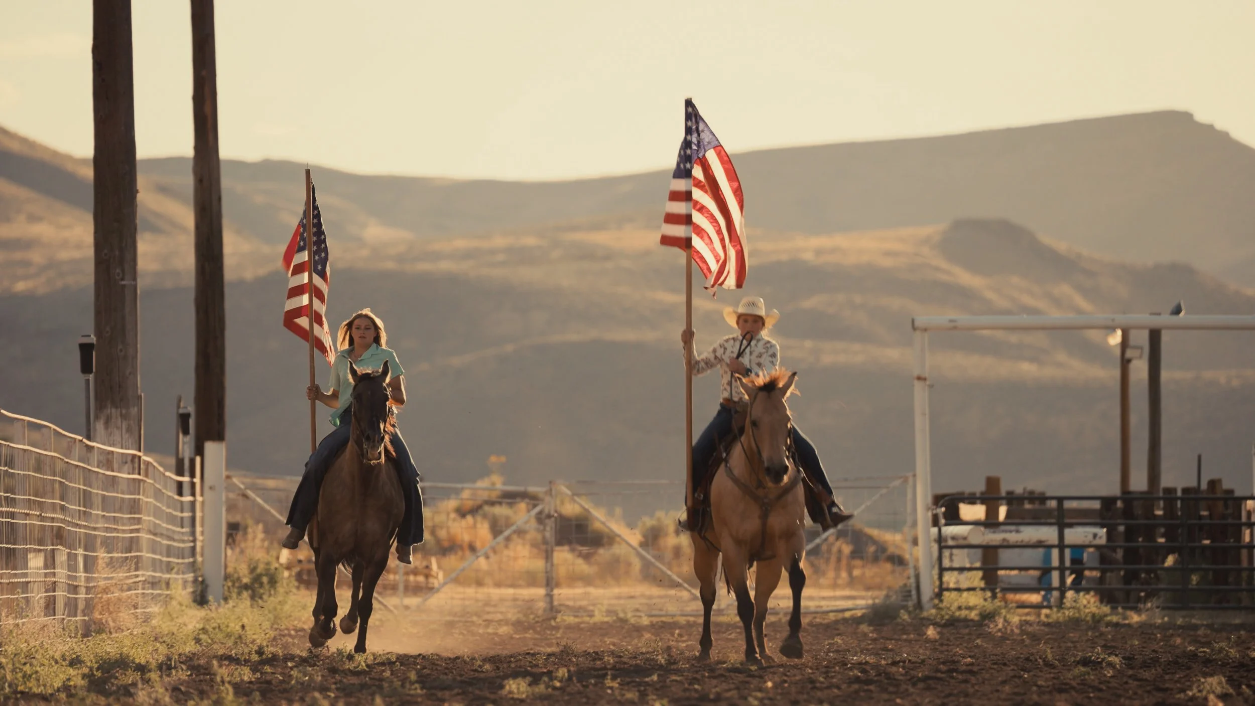Two young girls riding horses in a rodeo arena, each holding American flags. One girl is wearing a light green shirt, and the other is in a western hat and patterned shirt. The background features distant hills and a sunset sky.
