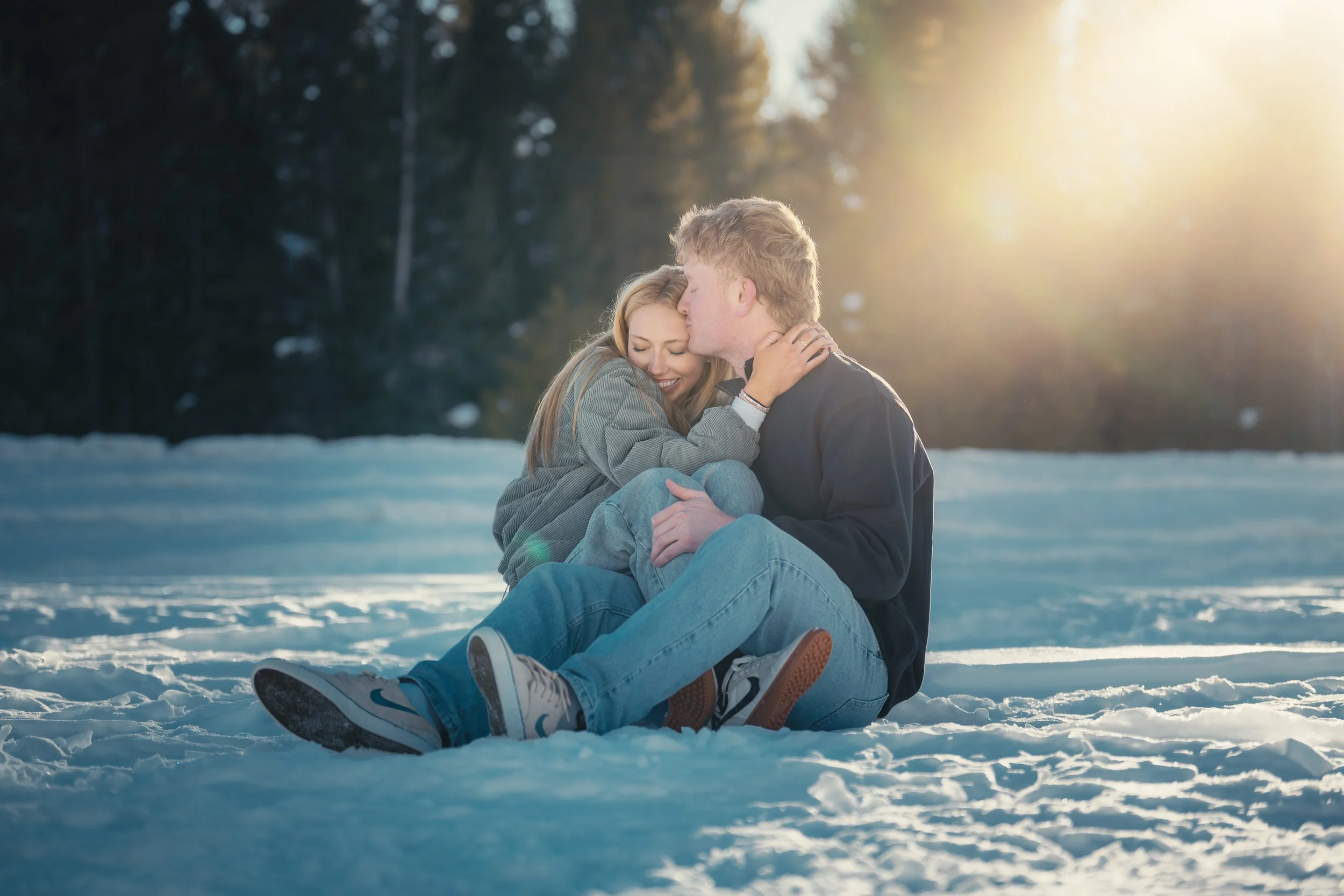 A young couple sitting on snow-covered ground outdoors during sunset, embracing each other with the man kissing the woman's forehead, surrounded by a forest of tall trees.