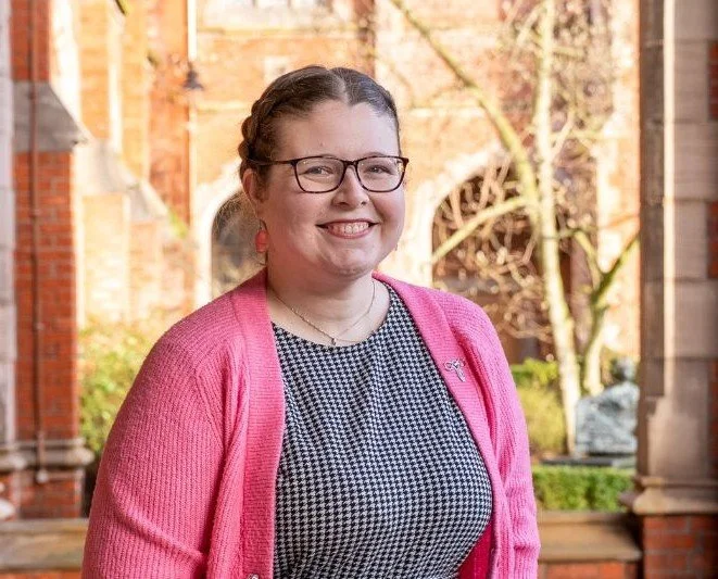 A portrait photograph of Dr. Robyn Atcheson, she is wearing a pink cardigan and a puppiestooth dress, and in the background the red and yellow Lanyon Building at Queen's can be seen.