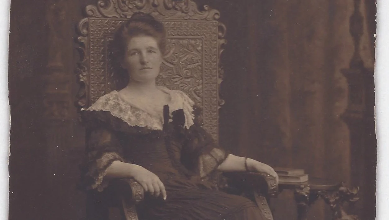 A black-and-white vintage photograph of a woman sitting in an ornate wooden chair with carved details, wearing a dark dress with lace accents, in front of a dark background with columns or decorative panels.