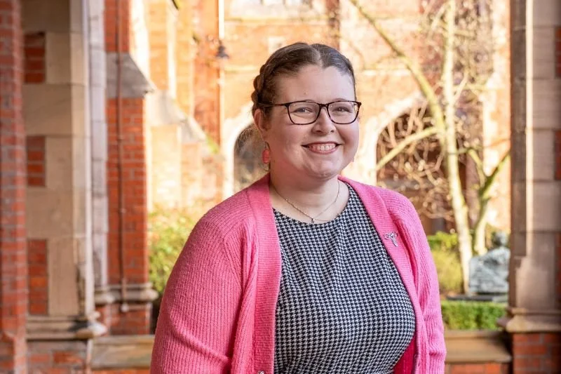 A portrait photograph of Dr Robyn Atcheson. She is pictured wearing a pink cardigan with Queen's University Belfast in the background.