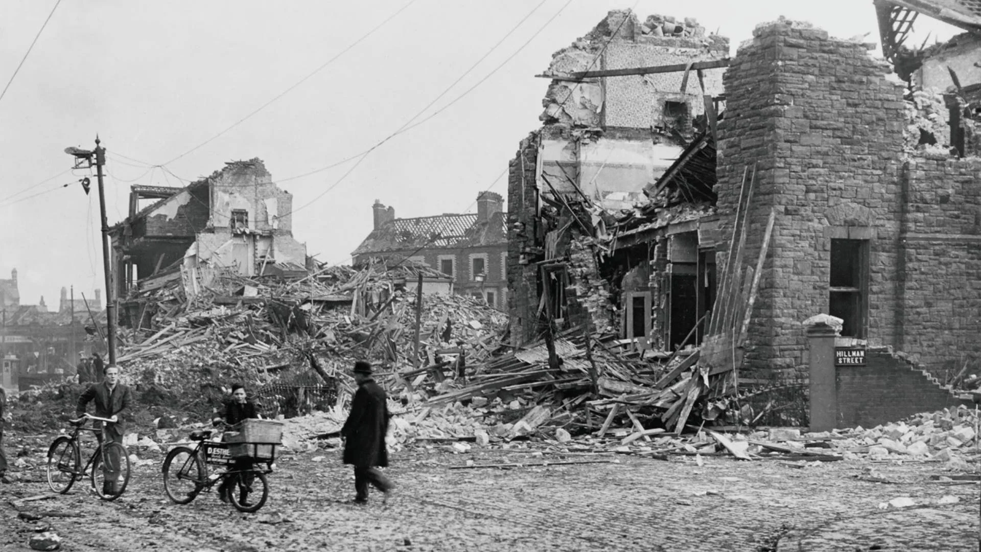 A photograph of the Antrim Road following the Easter Raid, part of the Belfast Blitz. In the Foreground a man and boy with bicycles are passed by a man in a long black coat, behind them the buildings are ruined, with rubble and timber strewn about.