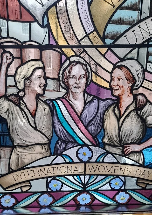 A stained glass window depicting three women celebrating International Women's Day, with one woman wearing a sash, surrounded by flowers and text.