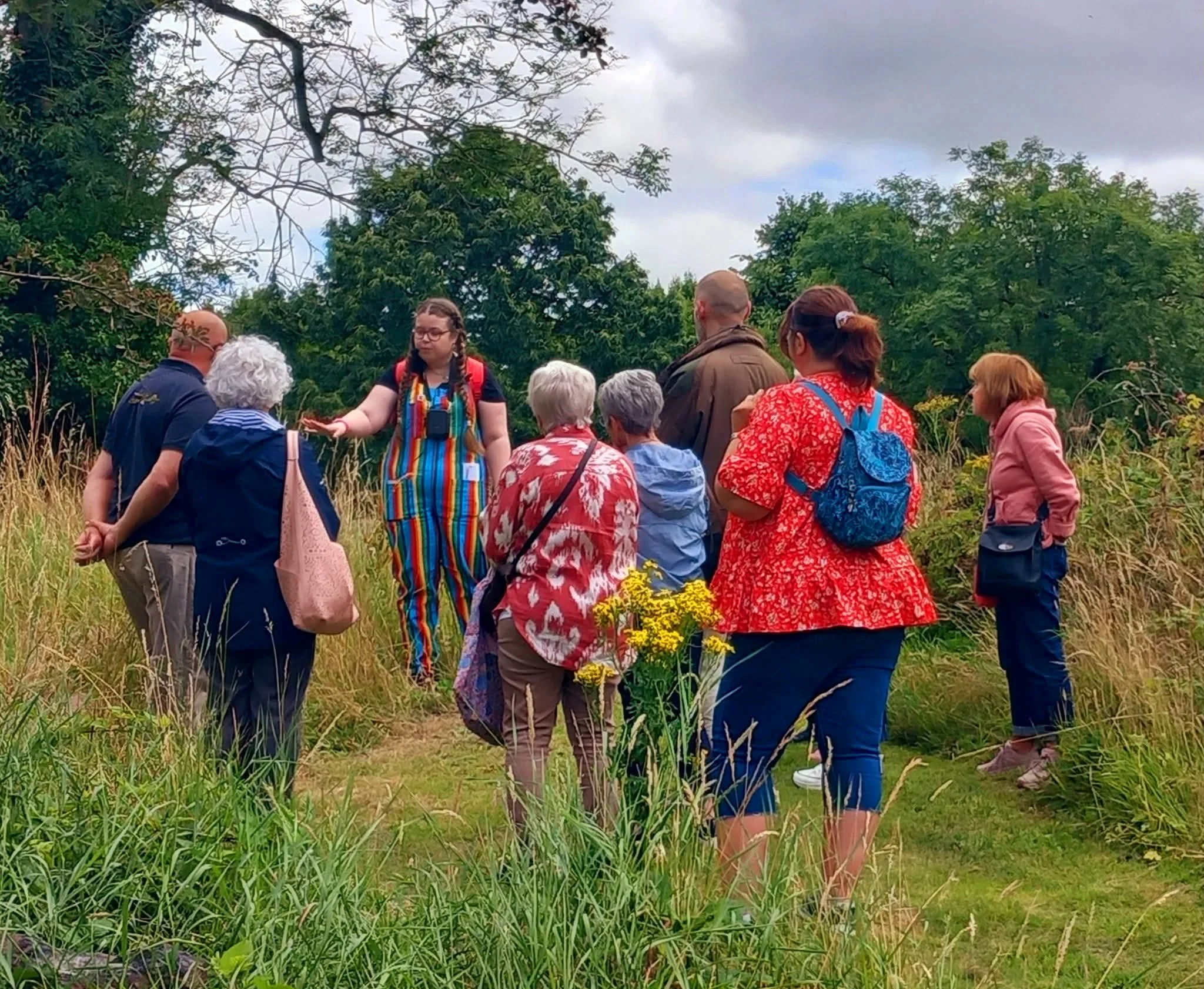 A group of diverse people, including women and men of various ages, gather outdoors in a lush garden or park, listening to a woman with braids and a striped apron who is gesturing as if explaining or guiding. The group appears engaged with tall green plants and trees in the background under a cloudy sky.
