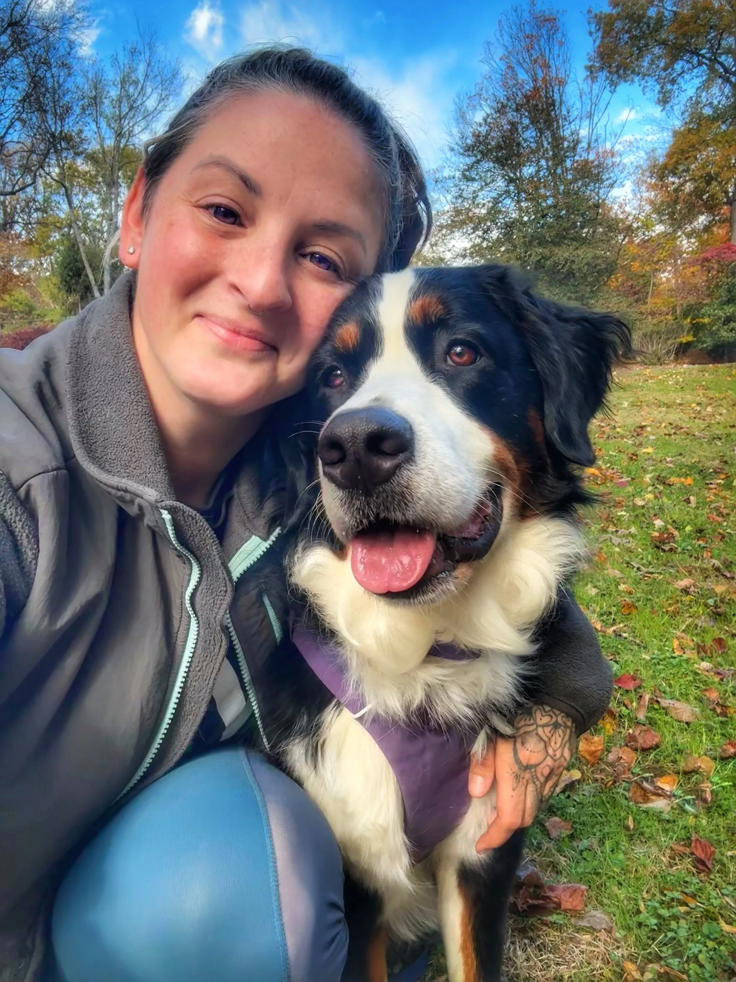 A white woman with brown hair and brown eyes smiles at the camera with her arm around a Bernese Mountain Dog.