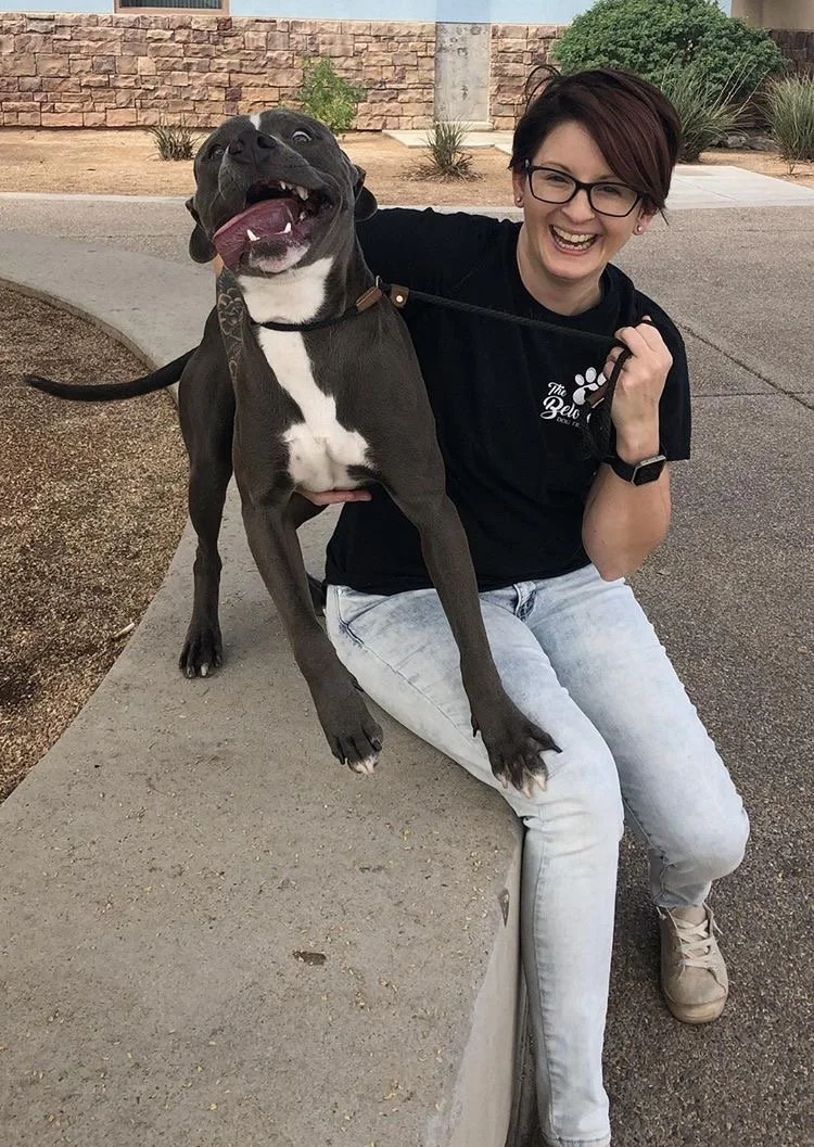 A gray and white pit bull with his tongue hanging out looking crazy stands on a ledge with a white woman with short reddish brown hair and who appears to be laughing.