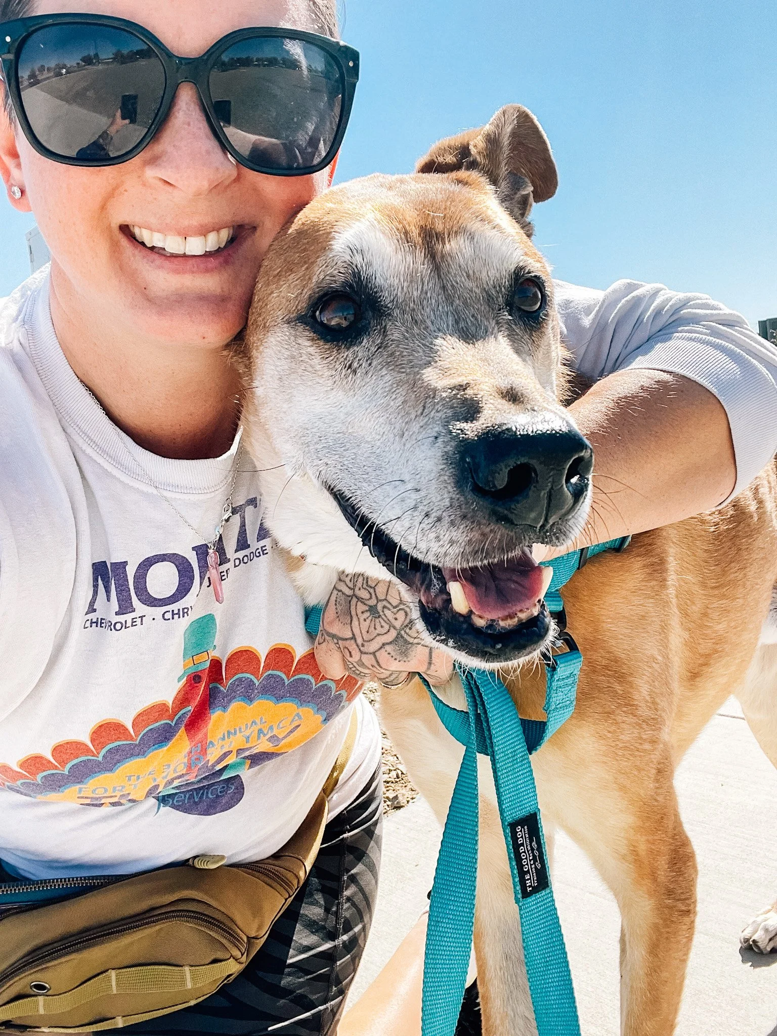 A white woman with big black sunglasses has her arm around a tall orange dog with a white face. They're both looking toward the camera and smiling.