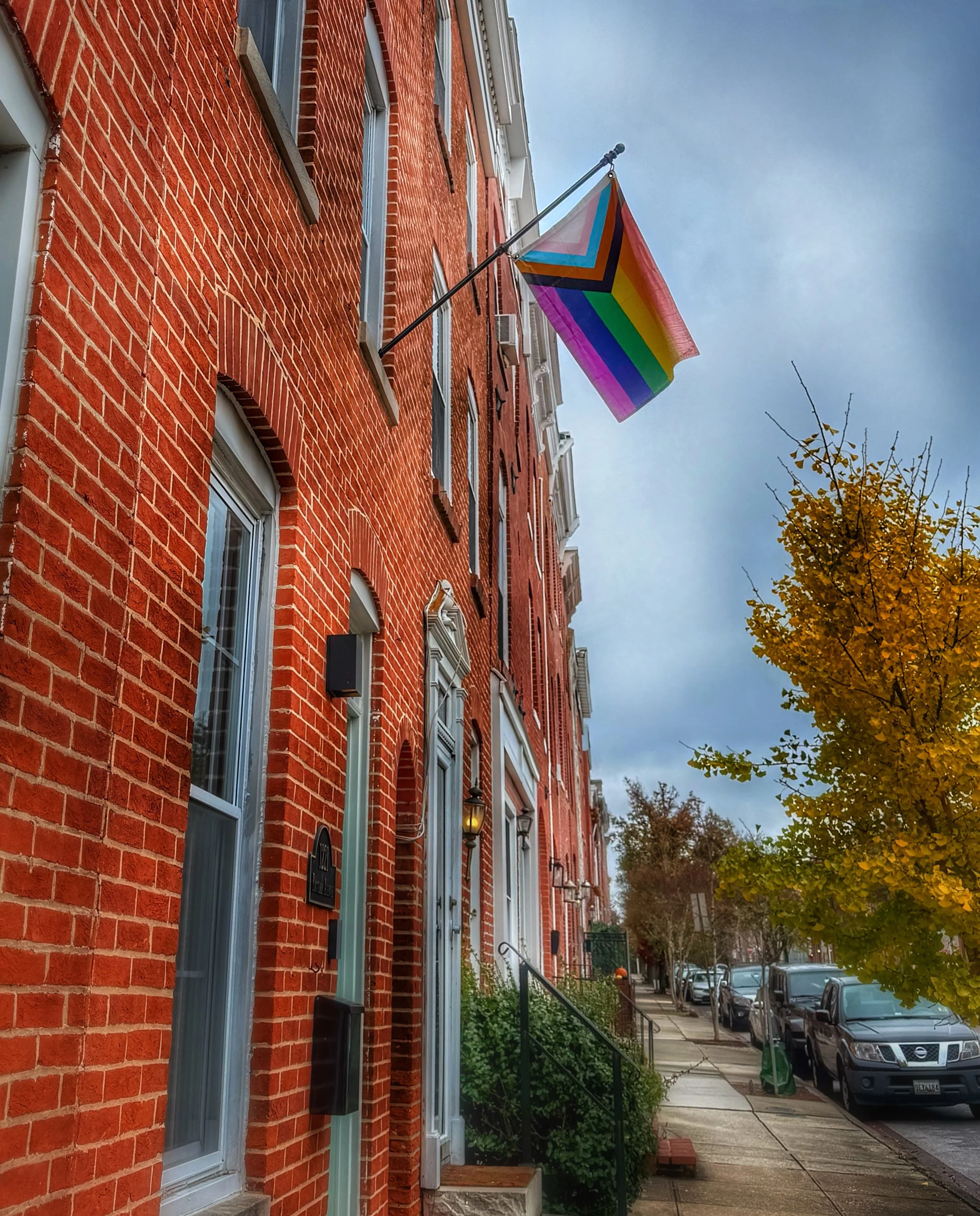 The camera looks along a row of a brick row homes with a flag pole bearing a progress pride flag.