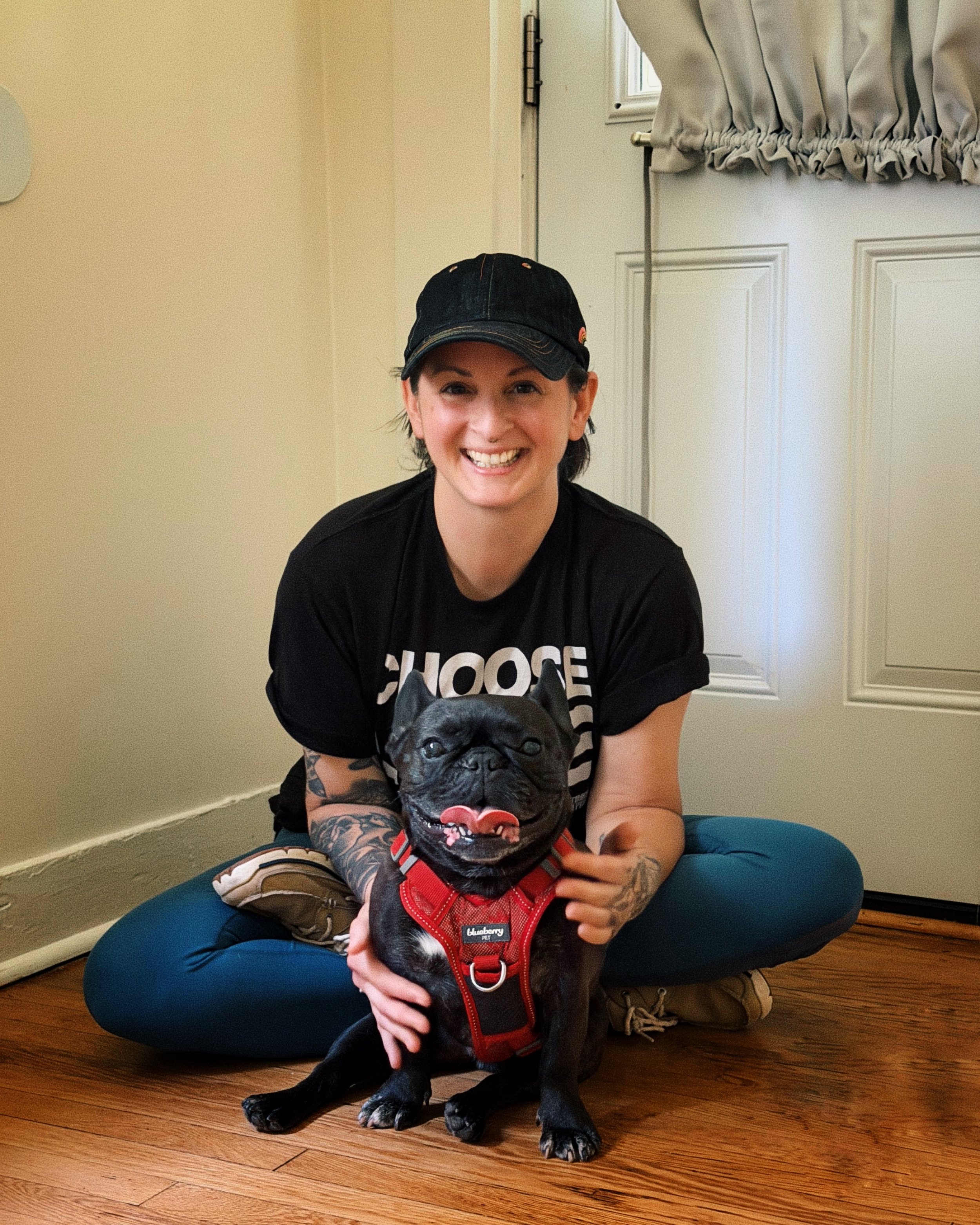 A white woman wearing a black hat and tshirt sits cross-legged on the floor with a black french bulldog in a red harness seated in front of her. Both are facing the camera and smiling.