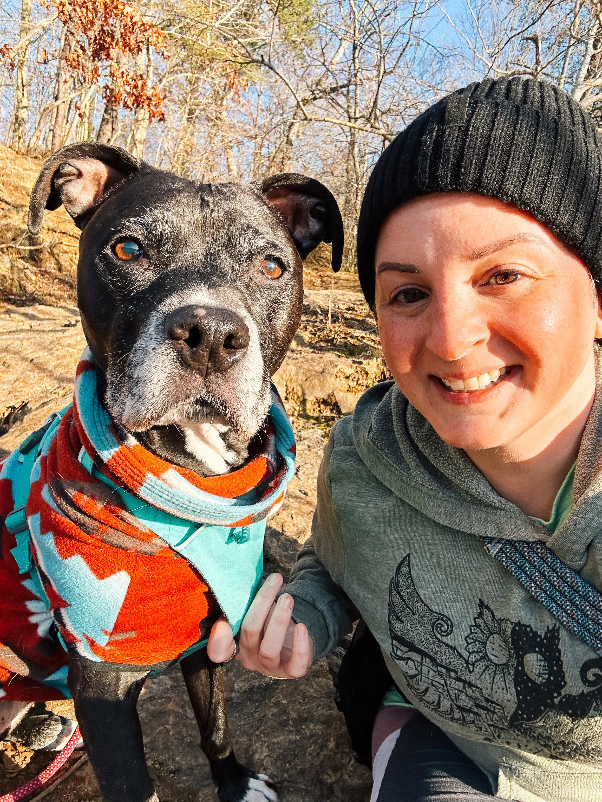 A black pit bull with a gray snout wearing a red and blue sweater looks at the camera with her ears perked. Next to her is a white woman in a black beanie and a green hoodie. They're in the woods.