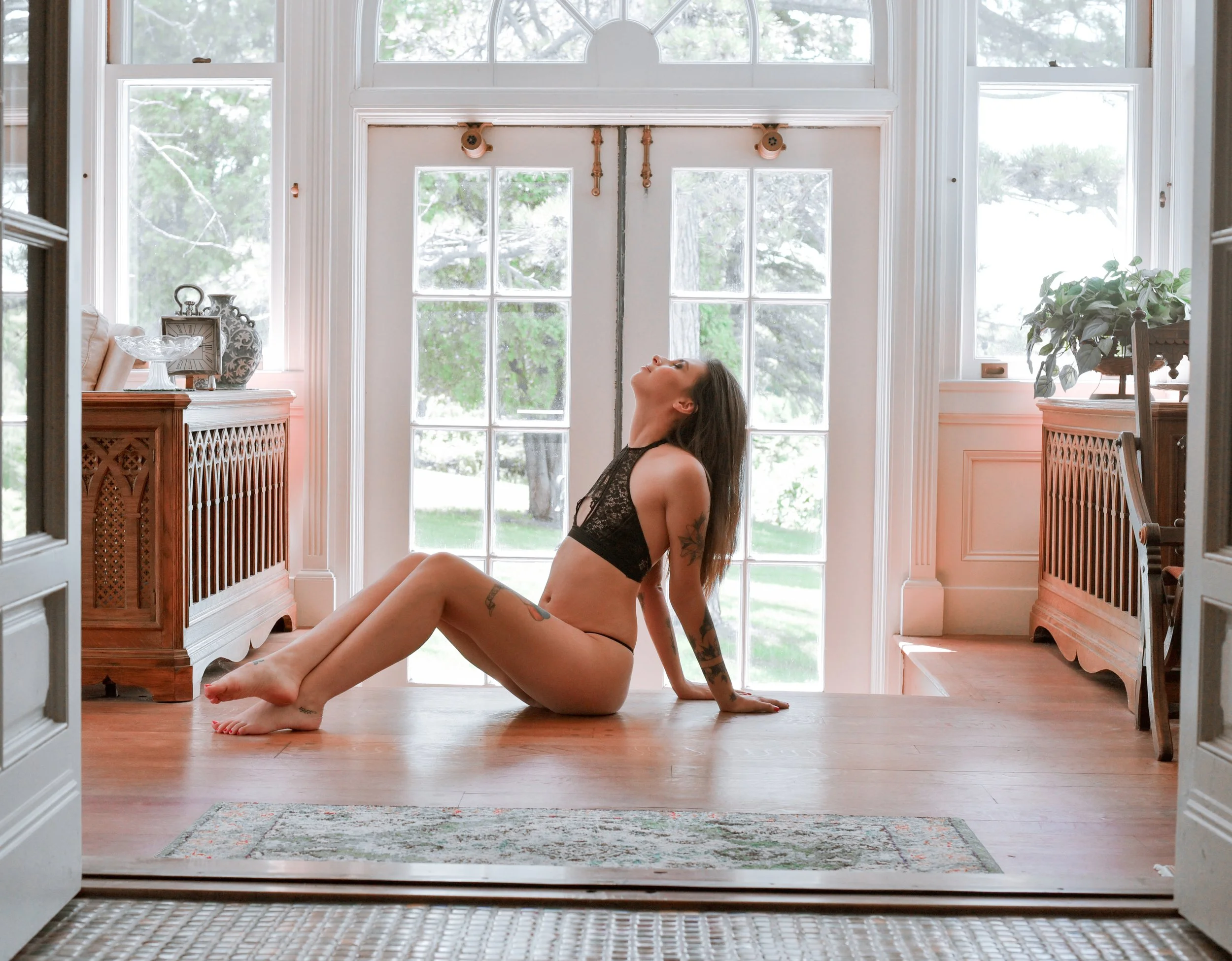 A woman with tattoos wearing black lace lingerie sitting on the wooden floor with a large window and green trees in the background.