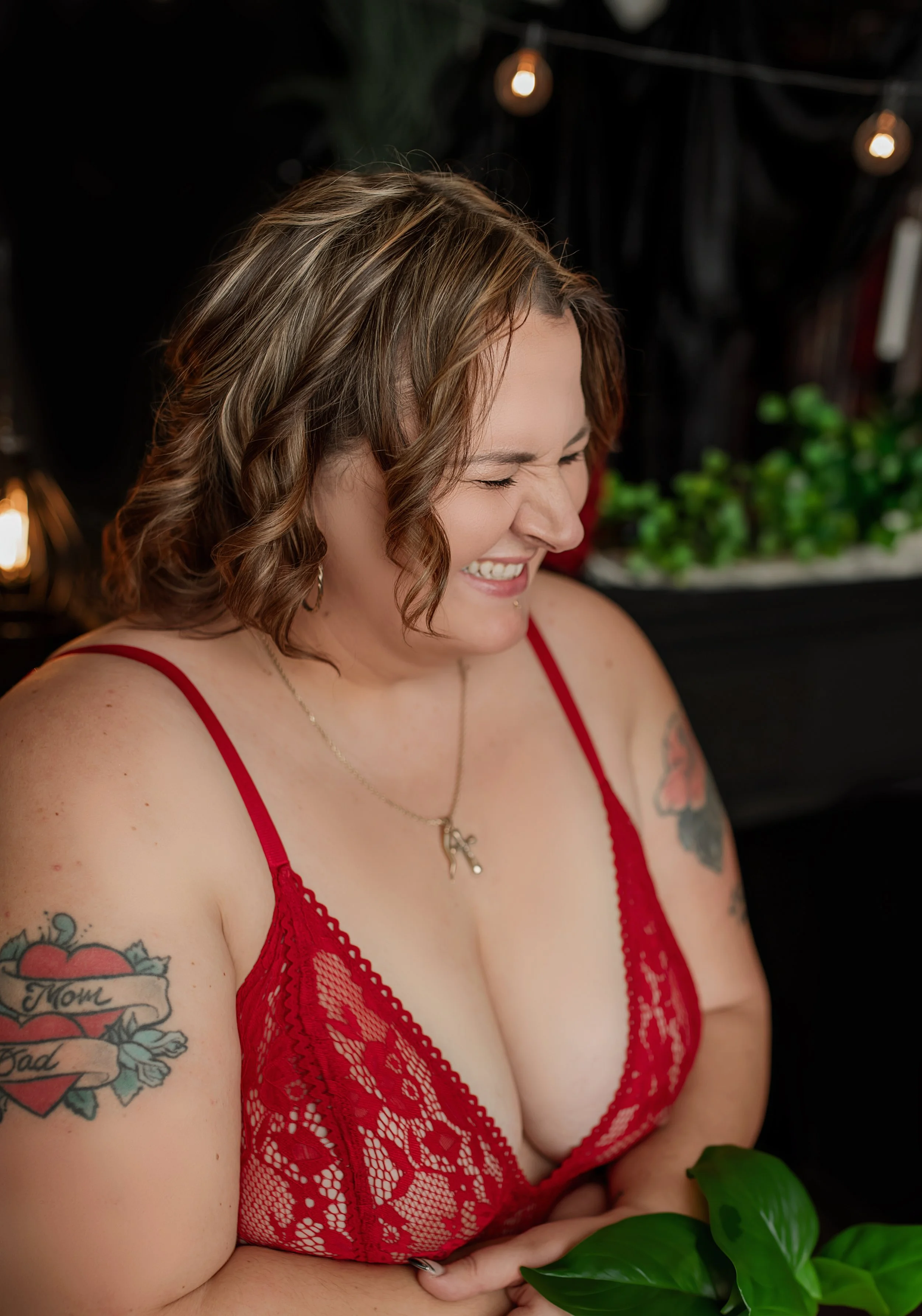 A woman with shoulder-length curly hair wearing a red lace dress, smiling with her eyes closed. She has tattoos on her arms, one reading "Mom" and "Dad" with a heart design. She is holding a green plant and wearing a necklace with a small key pendant