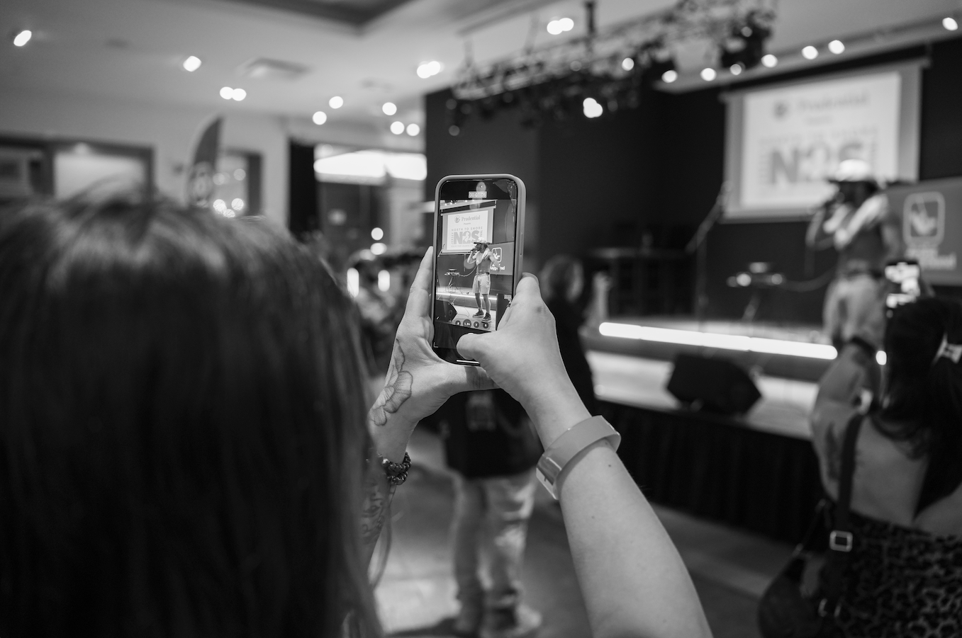 Person taking a photo of a performer on stage with a smartphone at an indoor event.