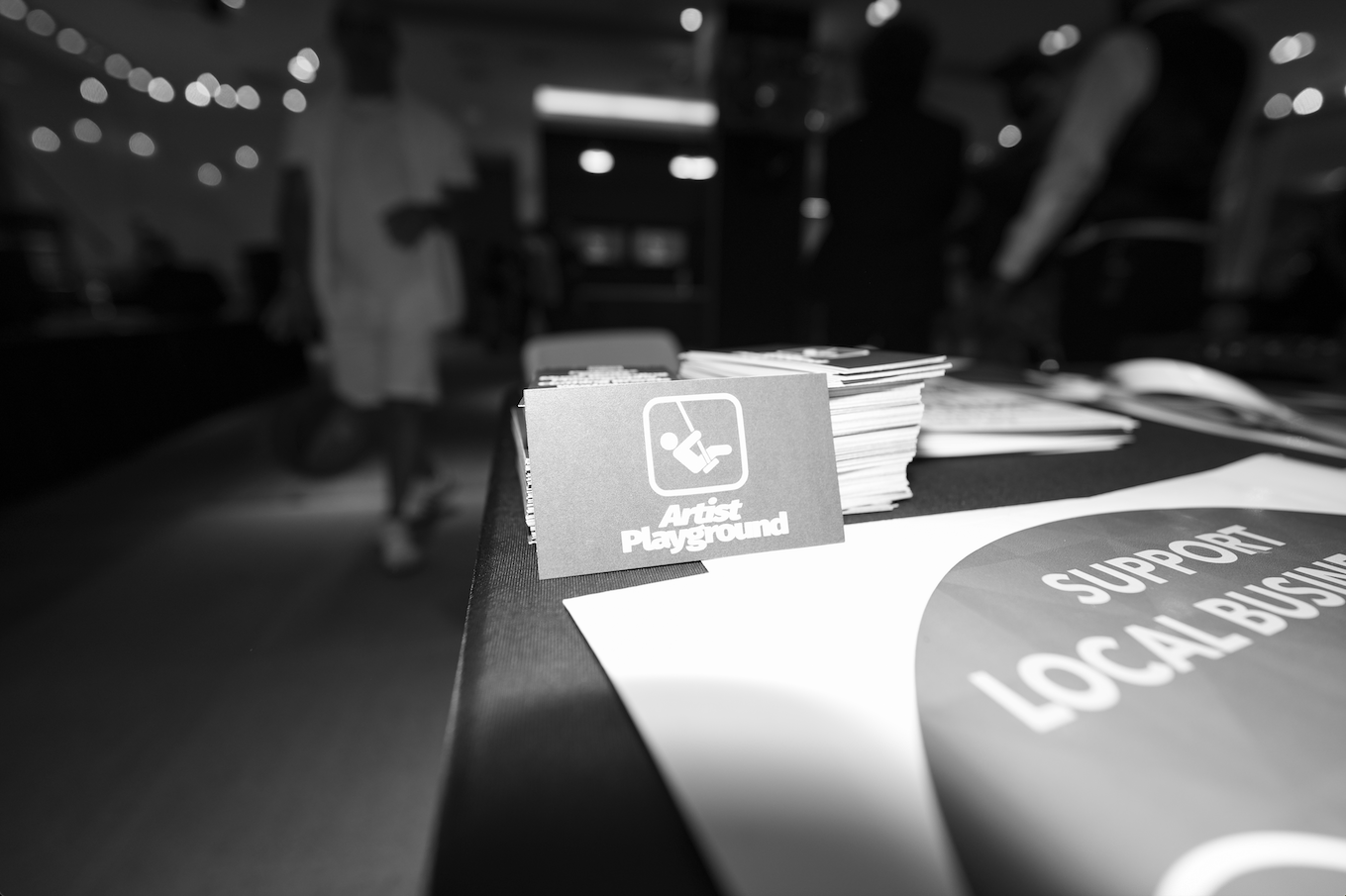 A table with informational and support materials for Art in the Playground, including a small sign with a wheelchair icon and the text "Artist Playground" in the foreground. Blurred background with people at an indoor event.