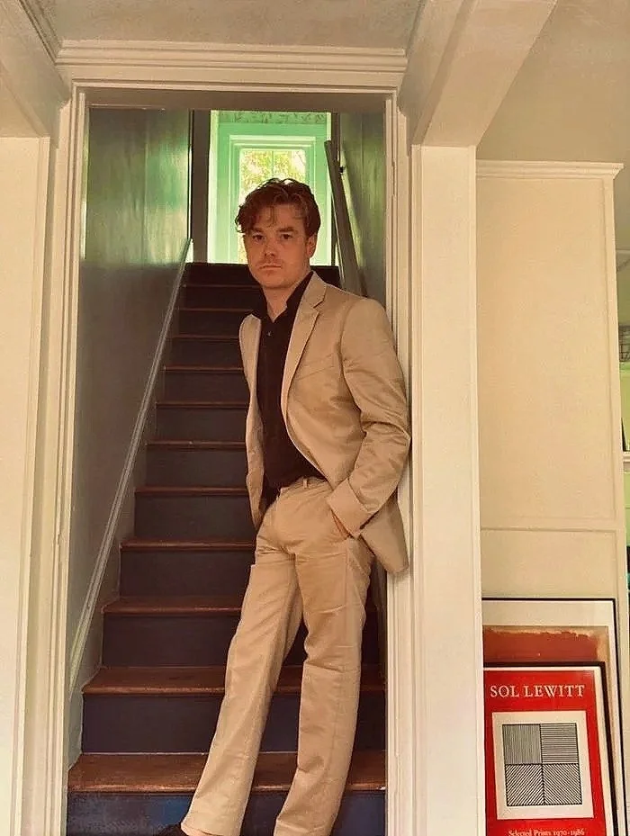 A young man in a beige suit and black shirt standing on a staircase in a house, with one hand in his pocket and looking at the camera.