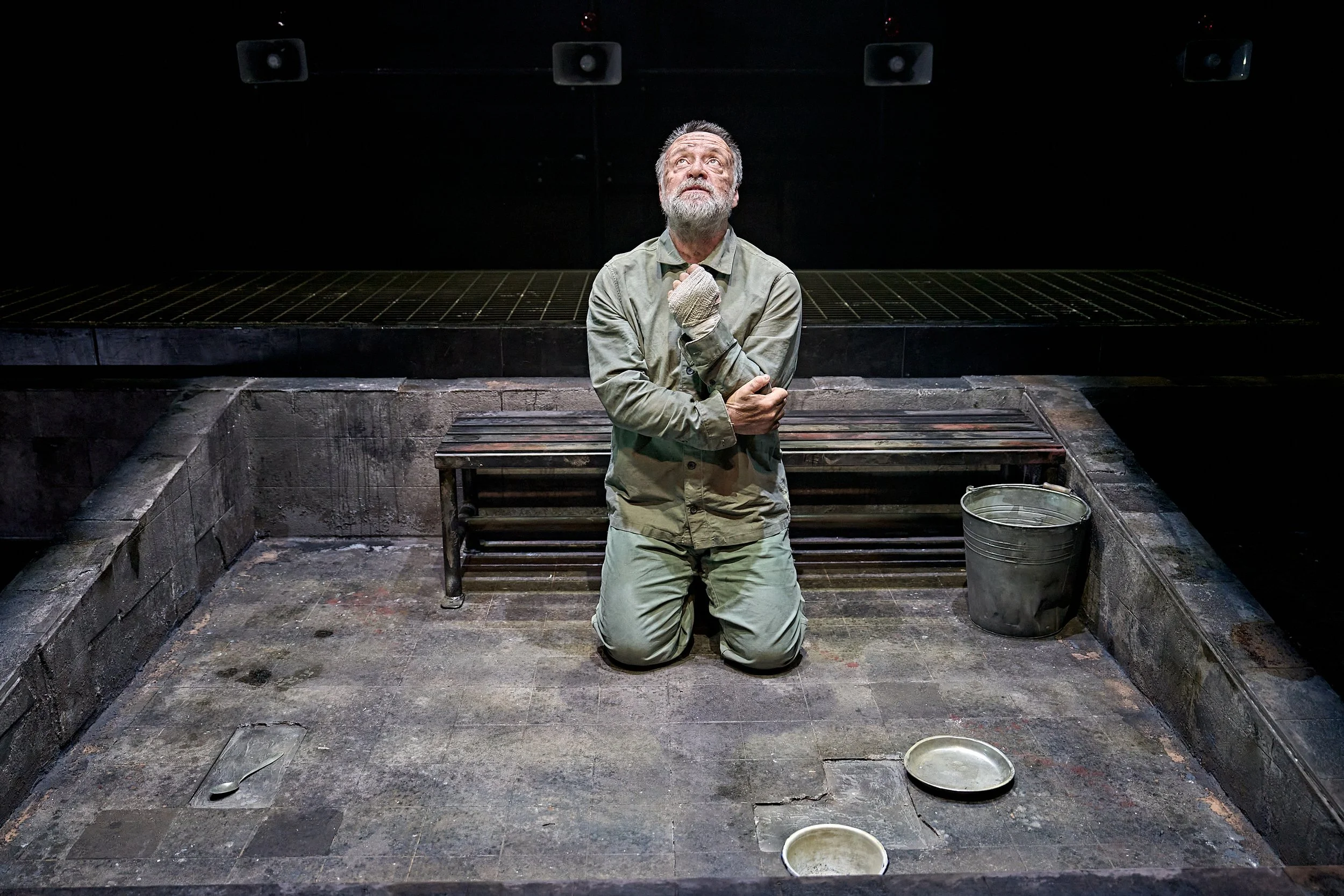 An elderly man with a beard, dressed in beige clothing and kneeling on a worn tiled floor, is holding his chest and looking upwards. The setting appears to be a dark, empty room with a black backdrop, a bench, and a metal bucket beside him.