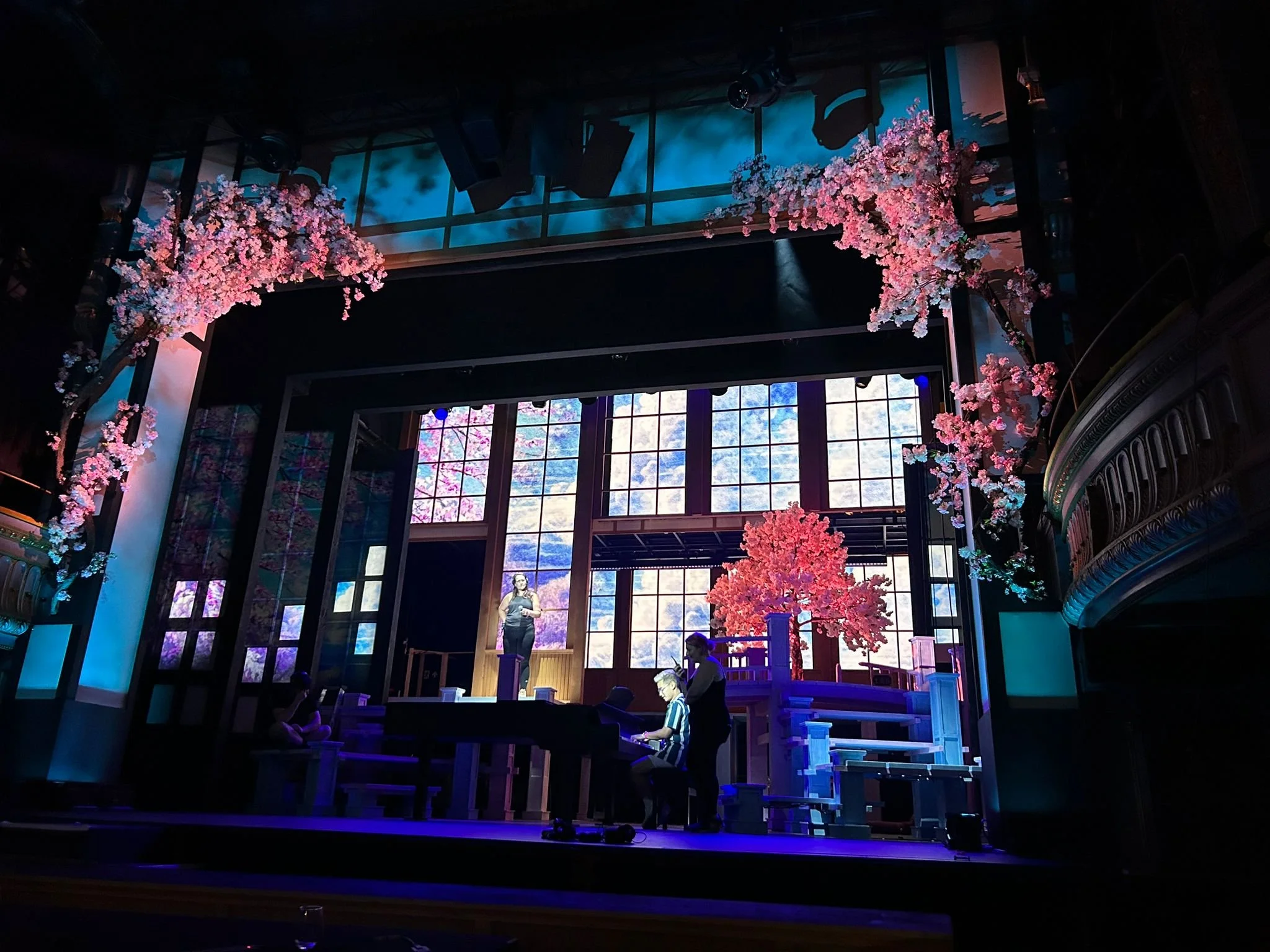 Stage set with pink cherry blossom trees, a woman on a balcony, a person playing piano, and bright background windows.