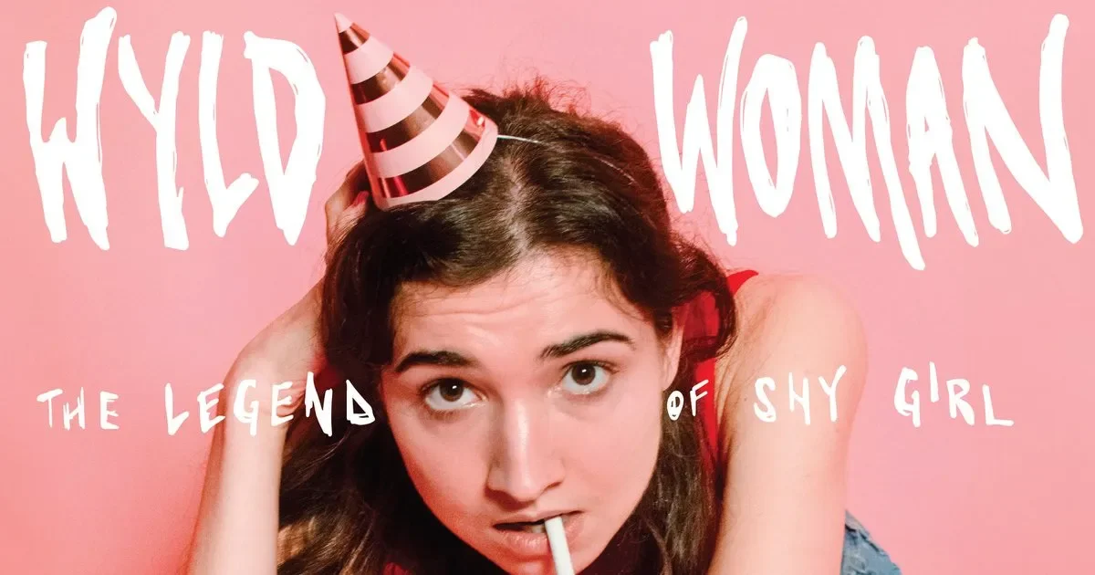 A young woman with long dark hair wearing a pink and white striped party hat and a red top. She has a lollipop in her mouth and is holding her head with one hand. The background is pink, with playful white text that reads "HYD WOMAN the legend of shy girl."