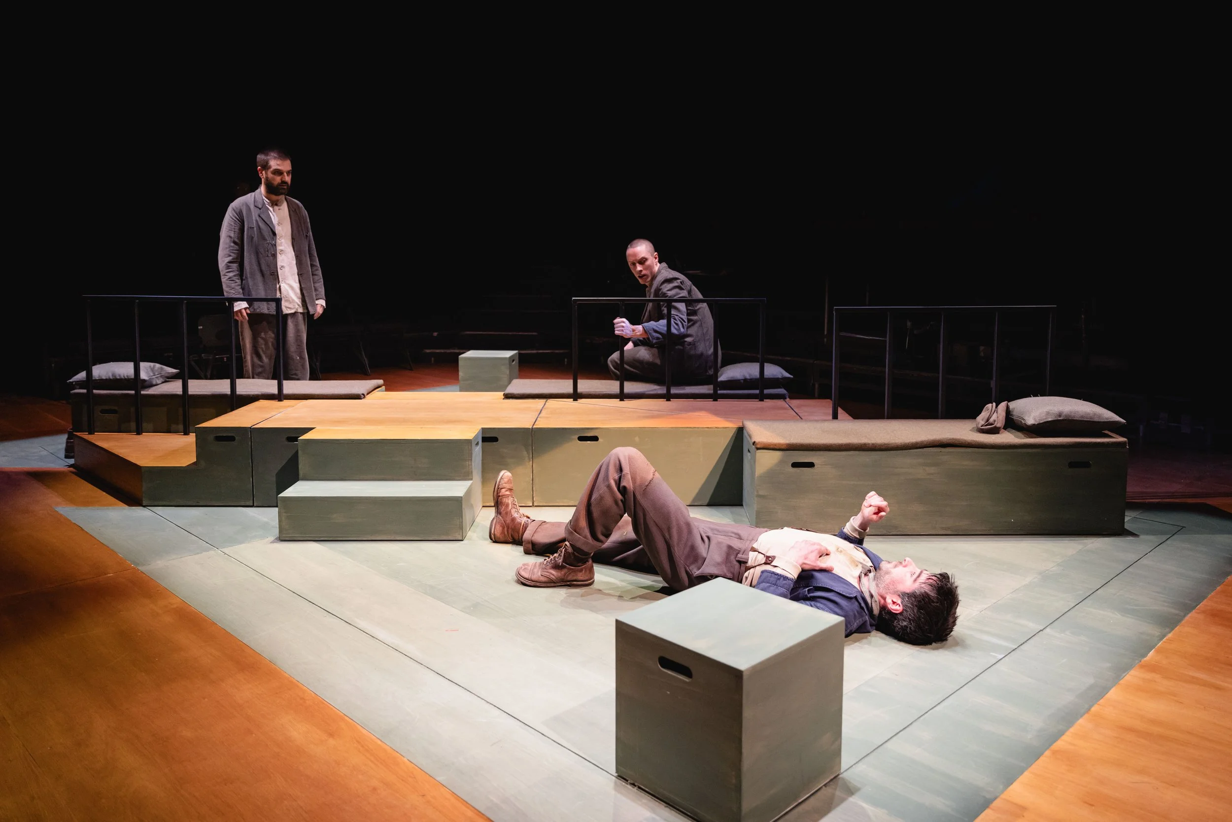 Three actors on a minimalist stage with wooden and green platform structures, pillows, and a dark backdrop, capturing a theatrical scene.