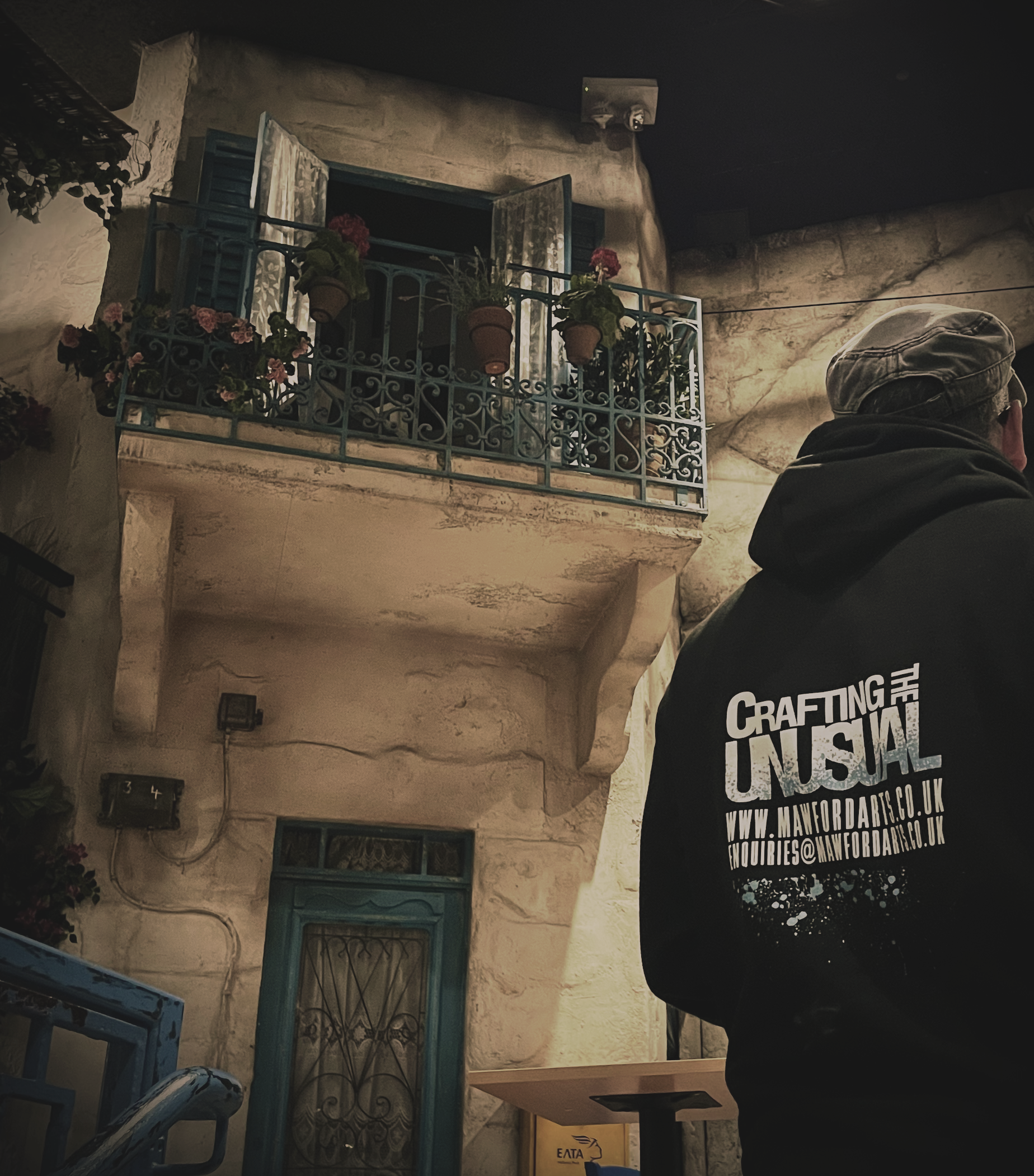 Person wearing a black hoodie with white text on the back, standing outside a building with an ornate balcony and potted plants, at night.