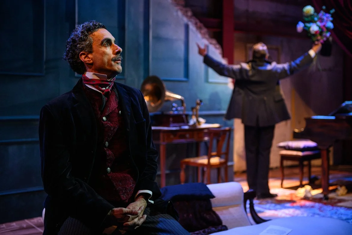 A man with dark curly hair and a beard, dressed in historical clothing, sitting in a room with a contemplative expression. In the background, a woman with short hair, dressed in a suit, is holding a bouquet of flowers and facing away with arms outstretched. The room has dark walls, wooden furniture, and vintage decor.