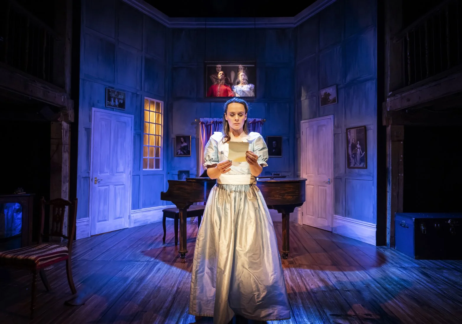 A woman in period costume standing at center stage in a theater or play setting, holding a letter or paper, with a set resembling a vintage room with blue walls, framed pictures, a grand piano, and curtains behind her.