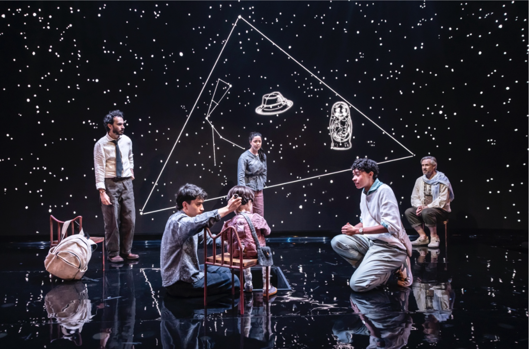 Group of six young people on stage, with a starry night sky and constellation drawings projected on a large screen behind them, engaging in a discussion or performance.