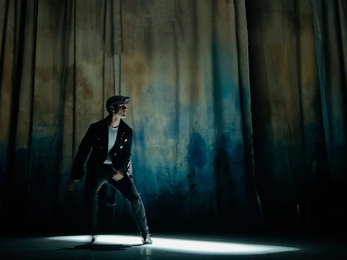A person performing a dance move in a dark setting with a single light source, wearing a black jacket and hat, against a backdrop of tall, dark curtains.