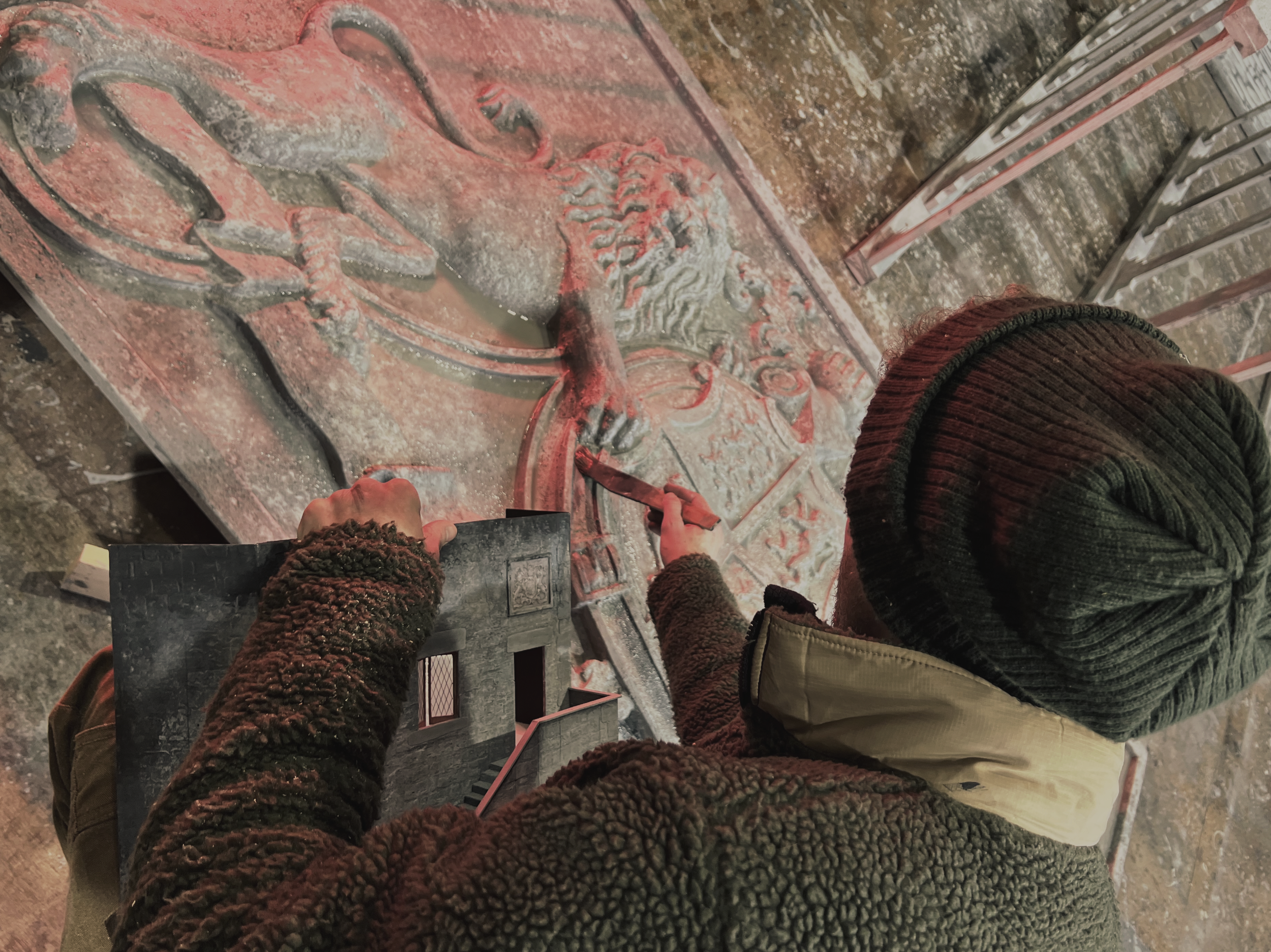 An artist carving a relief sculpture of a mythological scene on a stone slab. The artist is wearing a brown, fuzzy jacket and a green knit hat, and is using a chisel and hammer to work on the sculpture. The scene depicts a figure with a lion head and a shield, possibly a mythic or historical character.