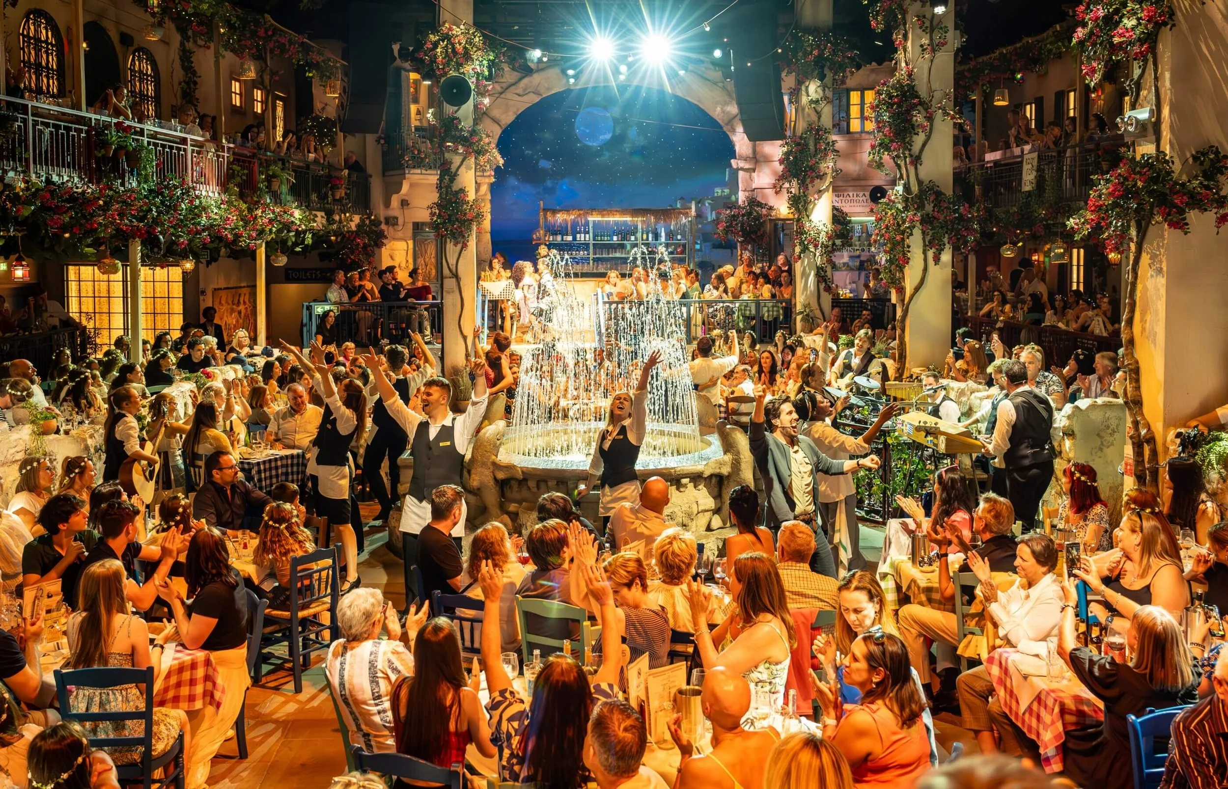 A lively indoor restaurant or bar with a central fountain, where many people are dining, drinking, and enjoying live music and dancing. The setting is decorated with flowers and has a festive atmosphere.