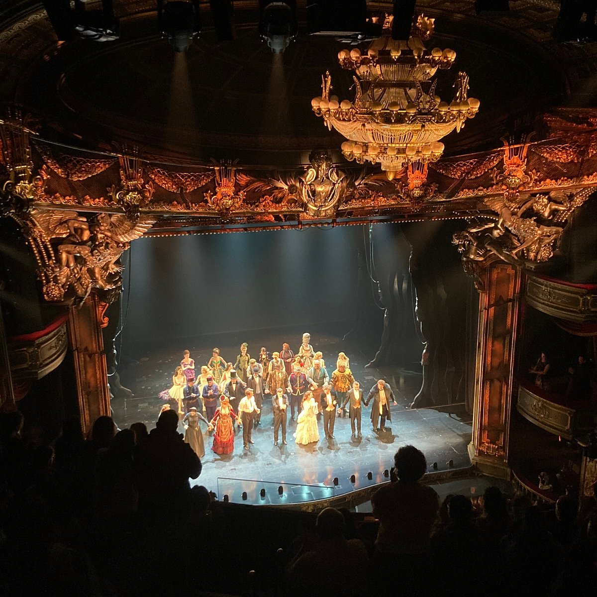 A theater stage with performers dressed in historical costumes, taking a bow after a performance. The theater is ornate with gold embellishments, a grand chandelier hanging from the ceiling, and an audience in the dark.