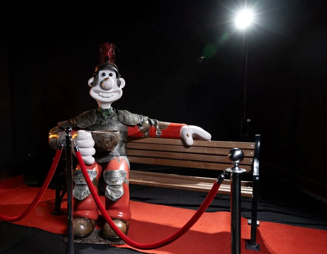 A life-sized statue of a cartoon character, possibly a warrior, is sitting on a wooden bench with a red carpet and velvet ropes around it, illuminated by a bright light in a dark room.