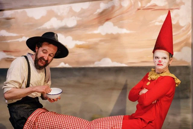 Clown with a tall red hat and white face paint sitting with arms crossed, next to a man dressed as a farmer holding a bowl, in front of a cloud-painted backdrop.