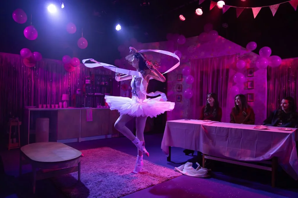 A ballerina performing a dance on a stage decorated with pink balloons and string curtains in a pink-themed room with three women watching and smiling.