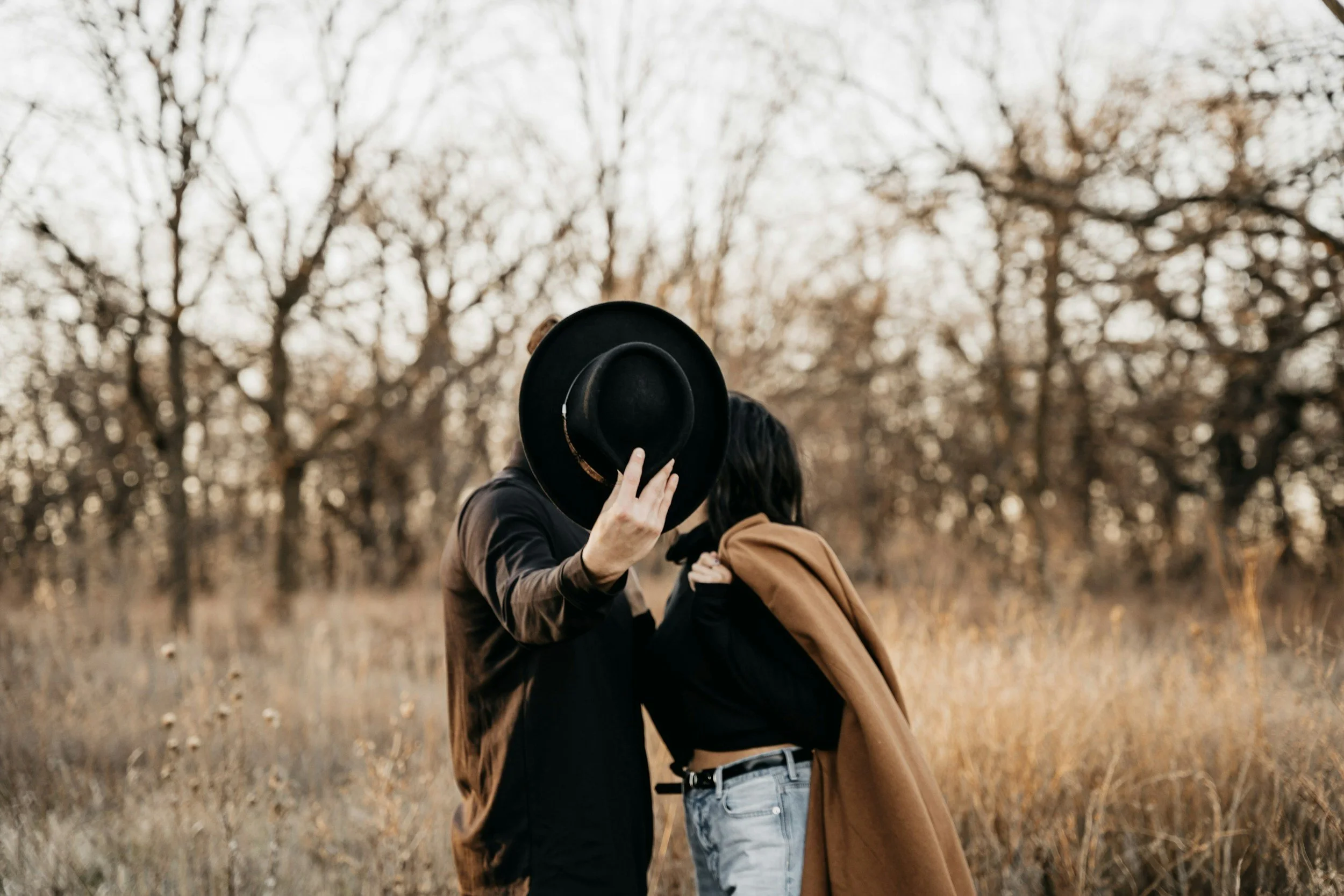 Two people standing in a field with trees in the background, one holding a black hat in front of their face, and the other with a brown coat draped over their shoulder.