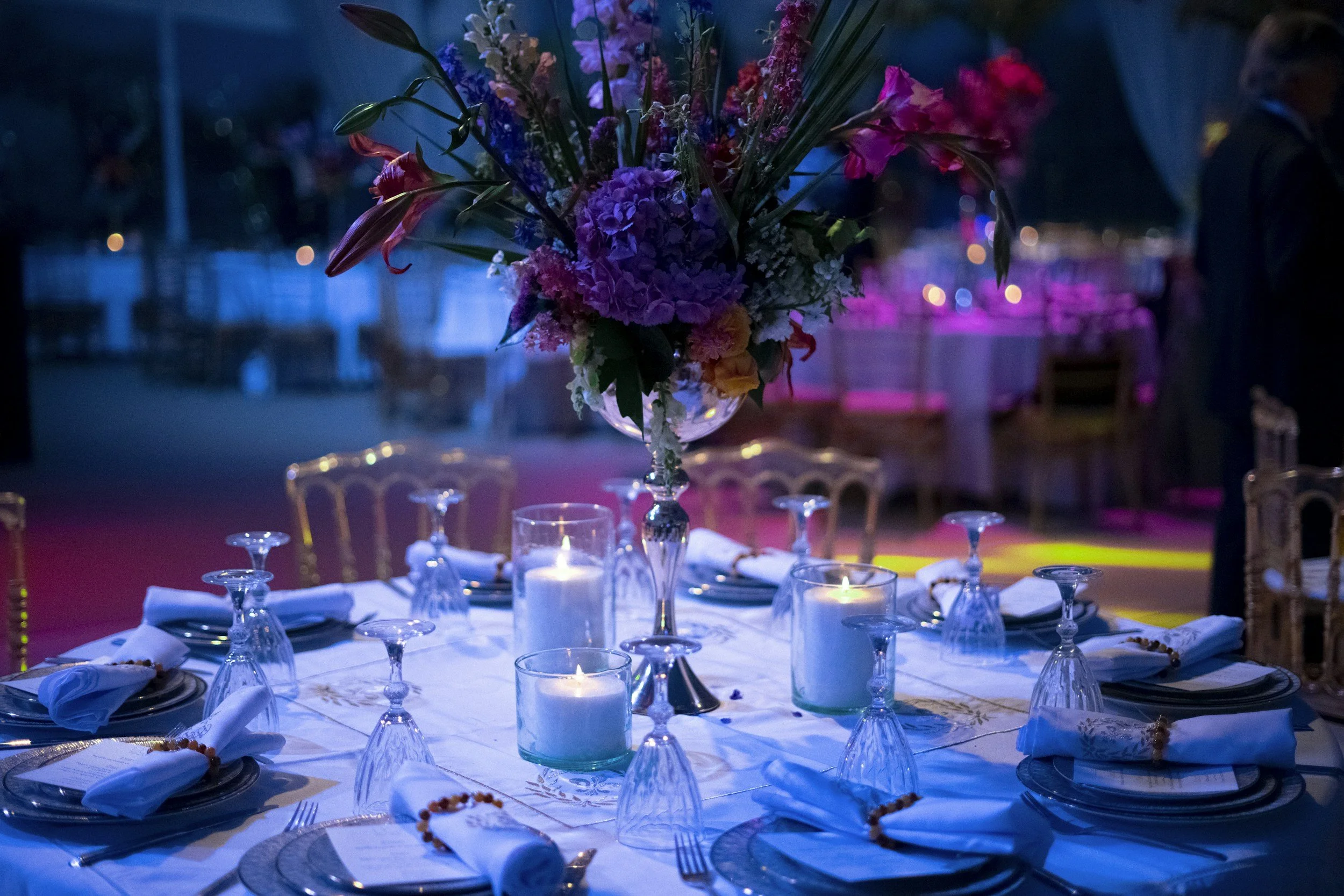 Round banquet table decorated with a large floral centerpiece, candles, place settings with napkins, glasses, and plates in a dimly lit event space with purple and blue lighting.