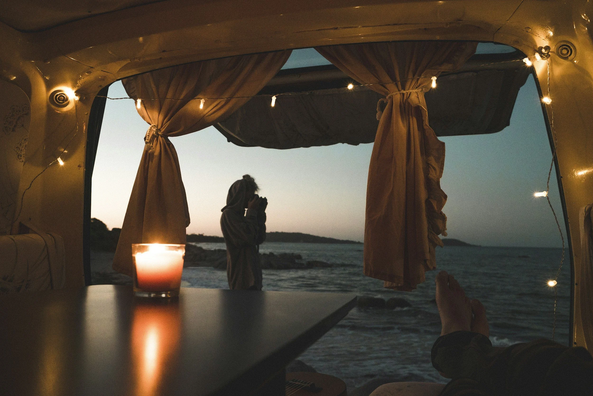Inside a camper van with a lit candle on the table, overlooking the beach at sunset. A person is sitting inside with their feet up, and another person is standing outside near the water, taking a photo.