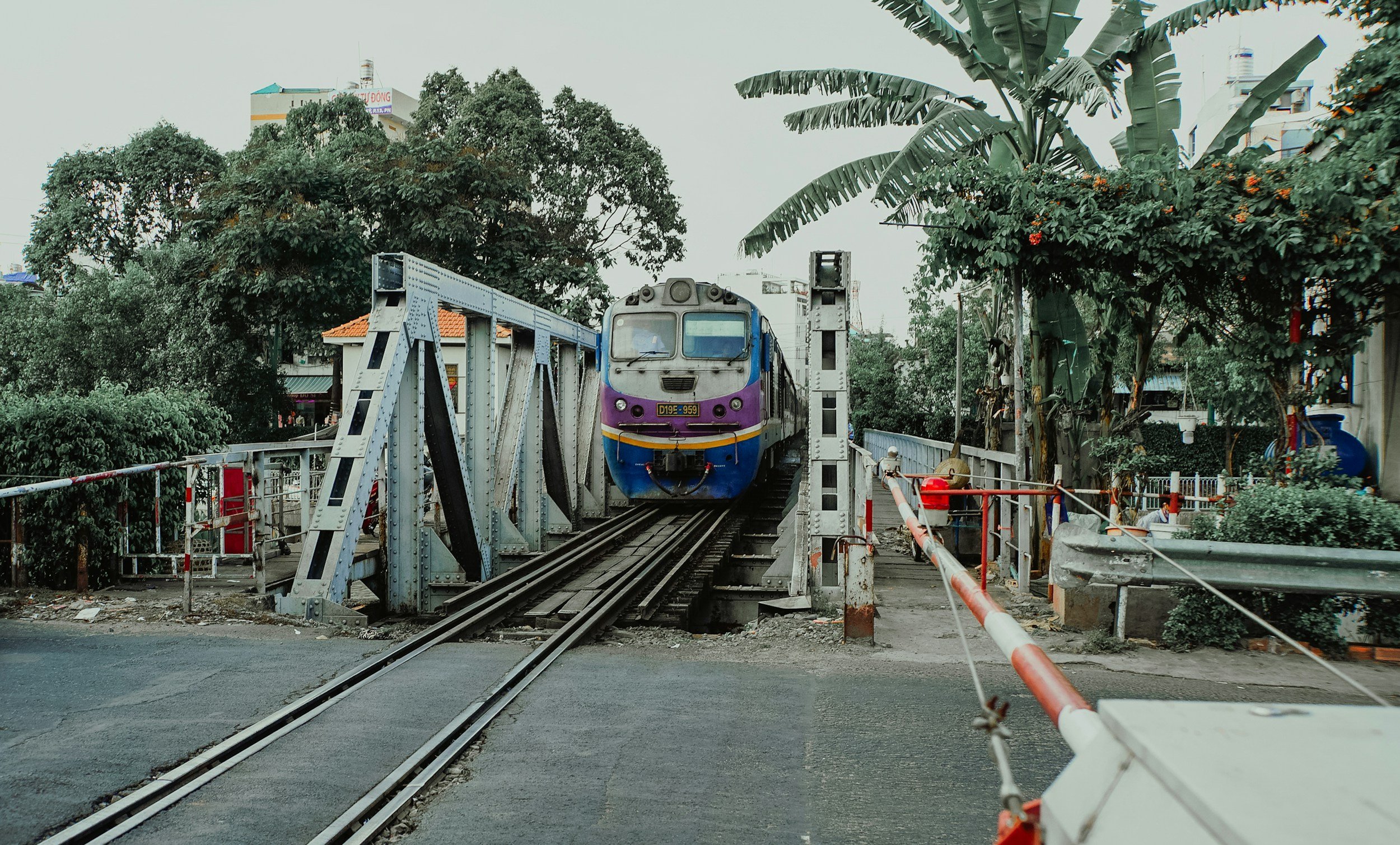 A train crossing a small metal bridge on railway tracks, with greenery and trees on both sides and buildings in the background.