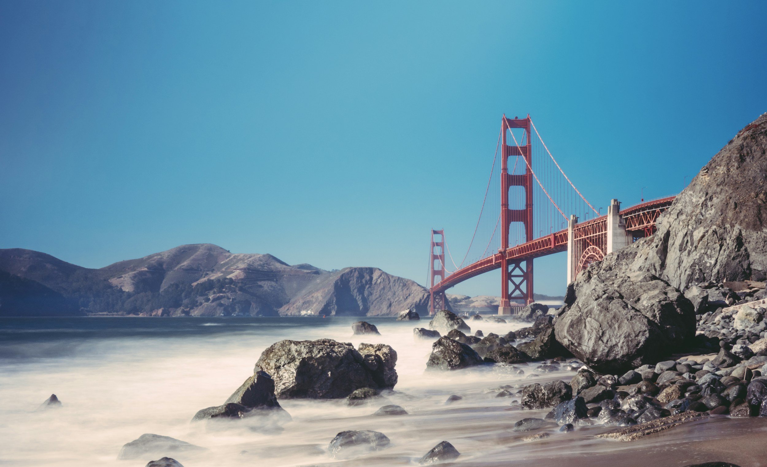 View of the Golden Gate Bridge in San Francisco, with rocks and shoreline in the foreground, water, and hills in the background under clear blue sky.
