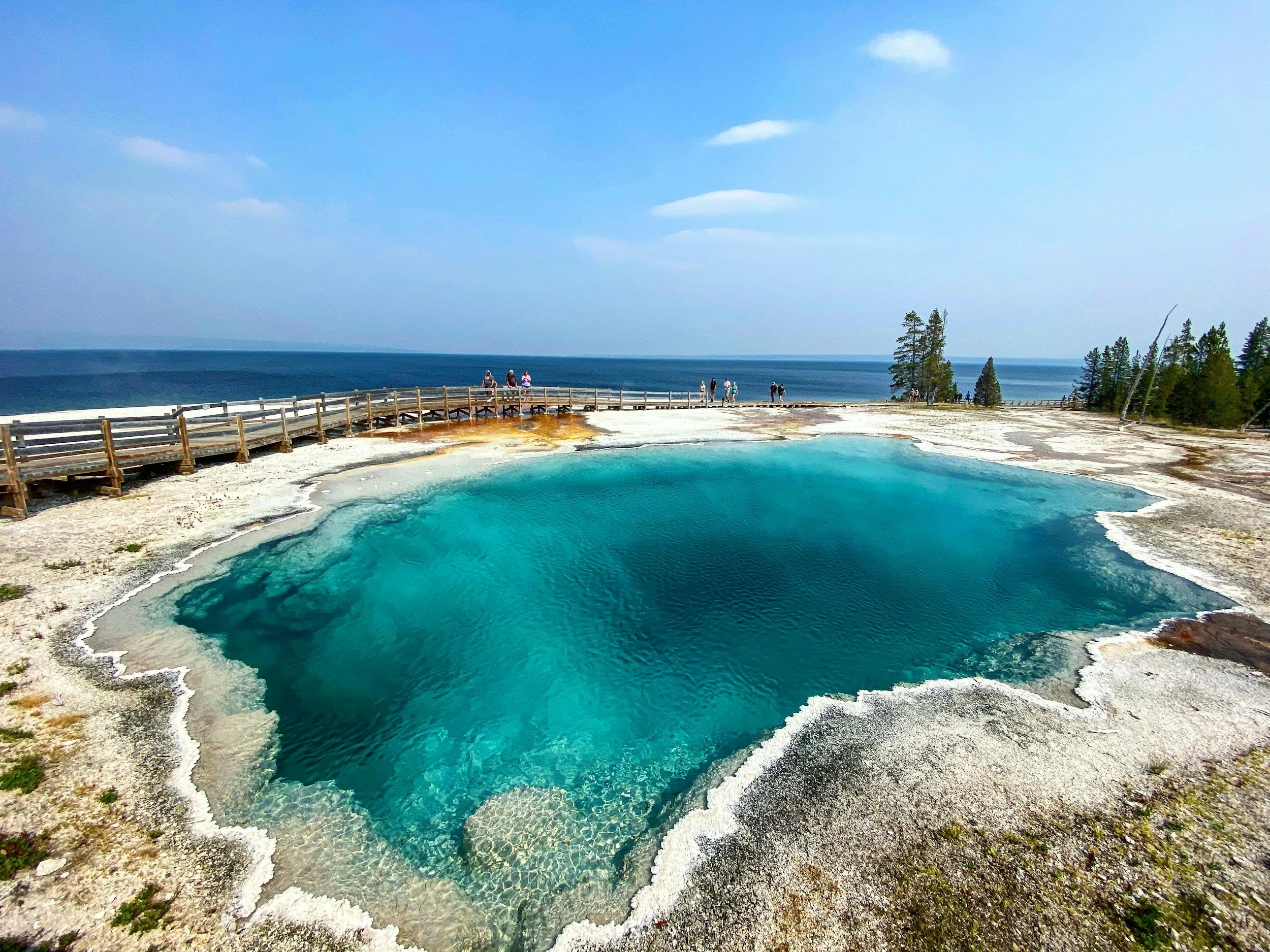 Hot spring with vibrant blue water, surrounded by mineral deposits, with a wooden boardwalk and visitors observing in the background, overlooking a large body of water under a partly cloudy sky.