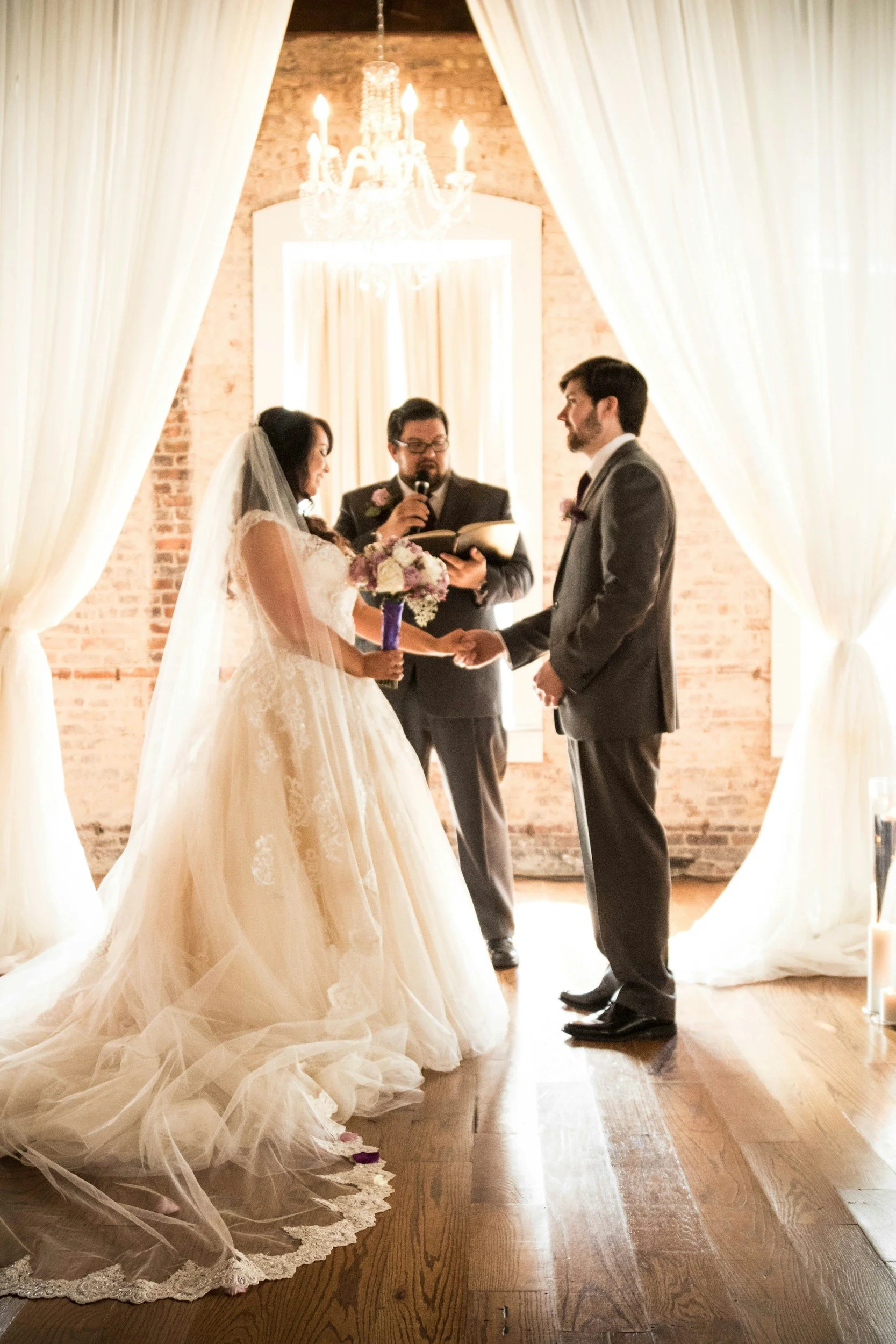 Wedding ceremony with bride and groom holding hands, officiant reading from a book, indoors with brick wall, chandelier, and white drapes.