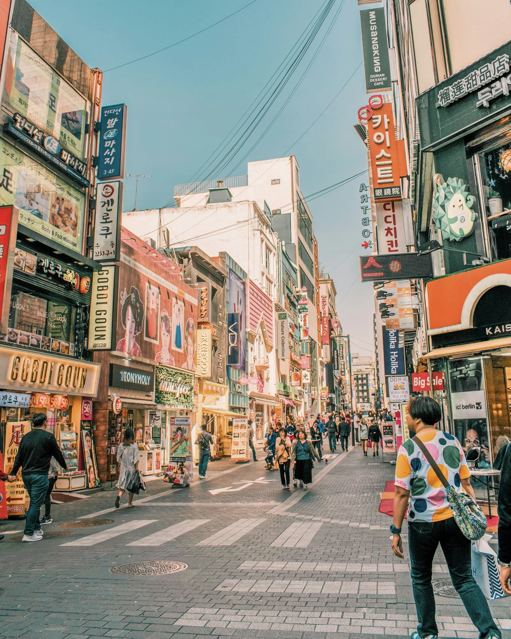 A bustling shopping street in Korea with colorful storefronts, neon signs, and pedestrians walking along the sidewalk during the day.