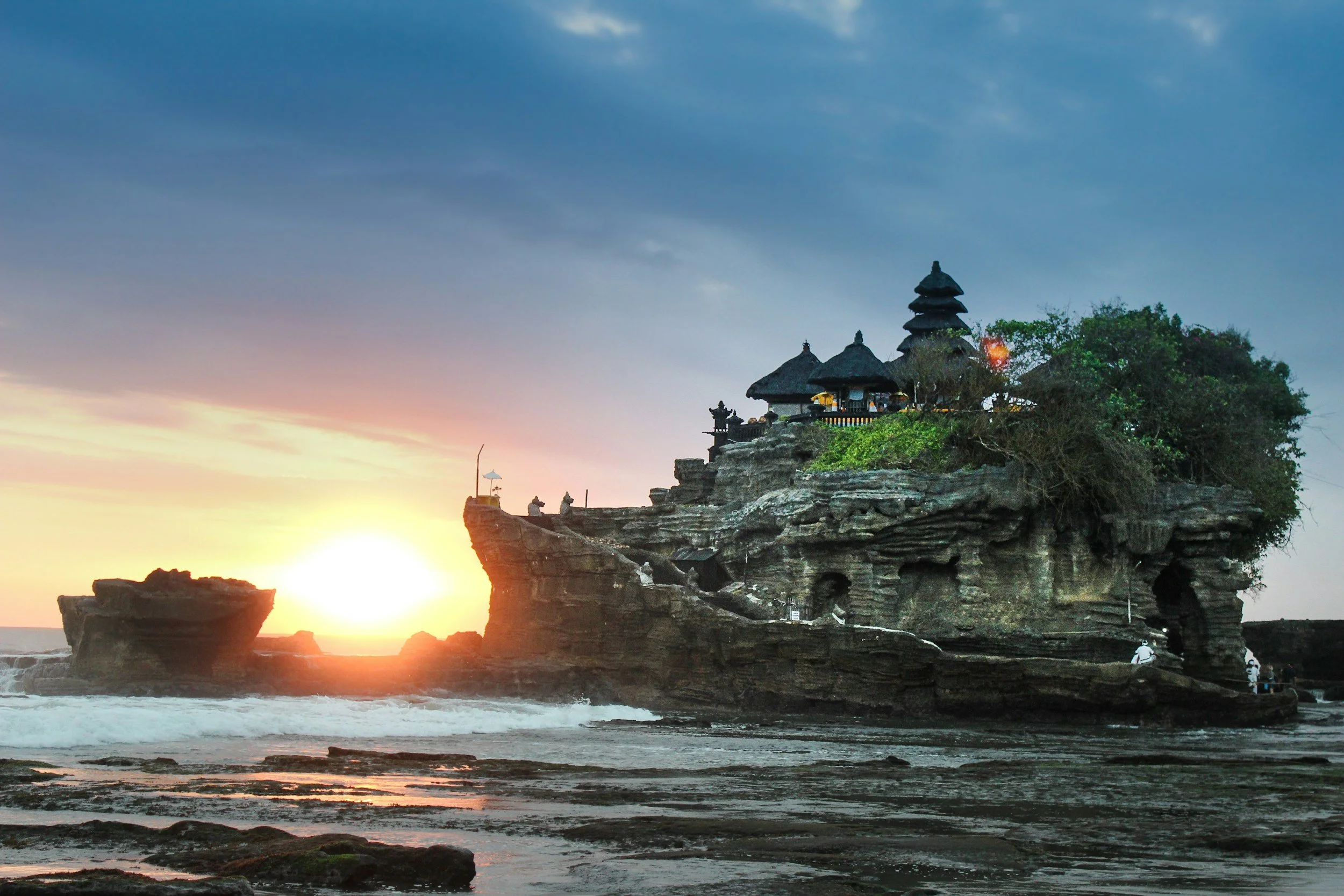 Sunset over a rocky coastal temple with traditional pagoda-style roofs, surrounded by trees and ocean waves.