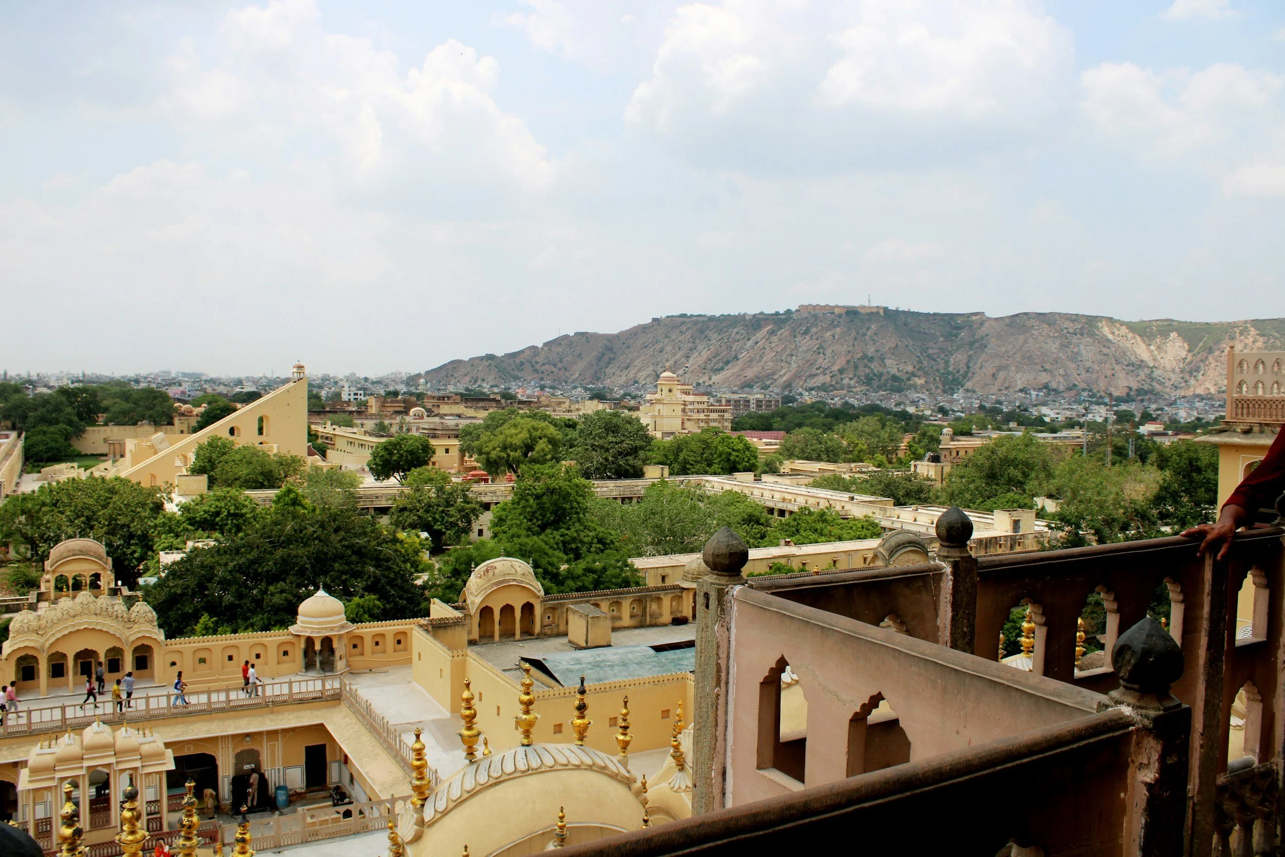 A panoramic view of a historic city with yellow sandstone buildings, lush green trees, and distant hills under a partly cloudy sky, as seen from a balcony.