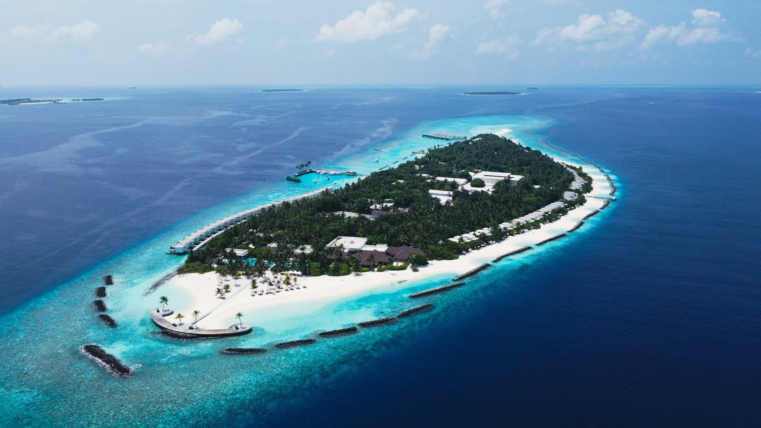 Aerial view of a tropical island resort with white sandy beaches, lined with palm trees, surrounded by turquoise waters and coral reefs.